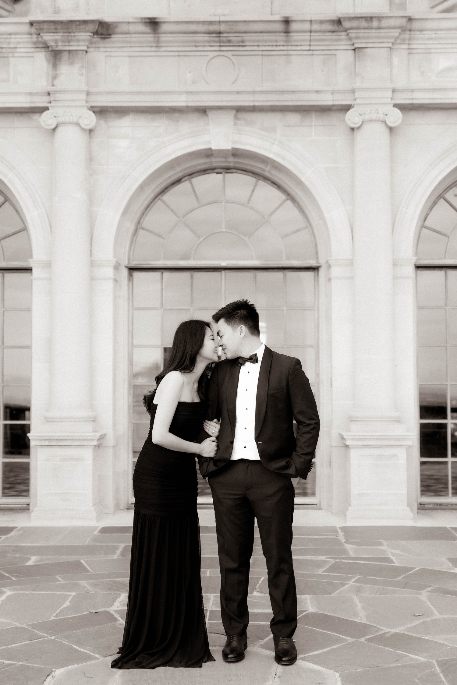 Couple in formal attire sharing a kiss in front of neoclassical arched building in black and white photo.