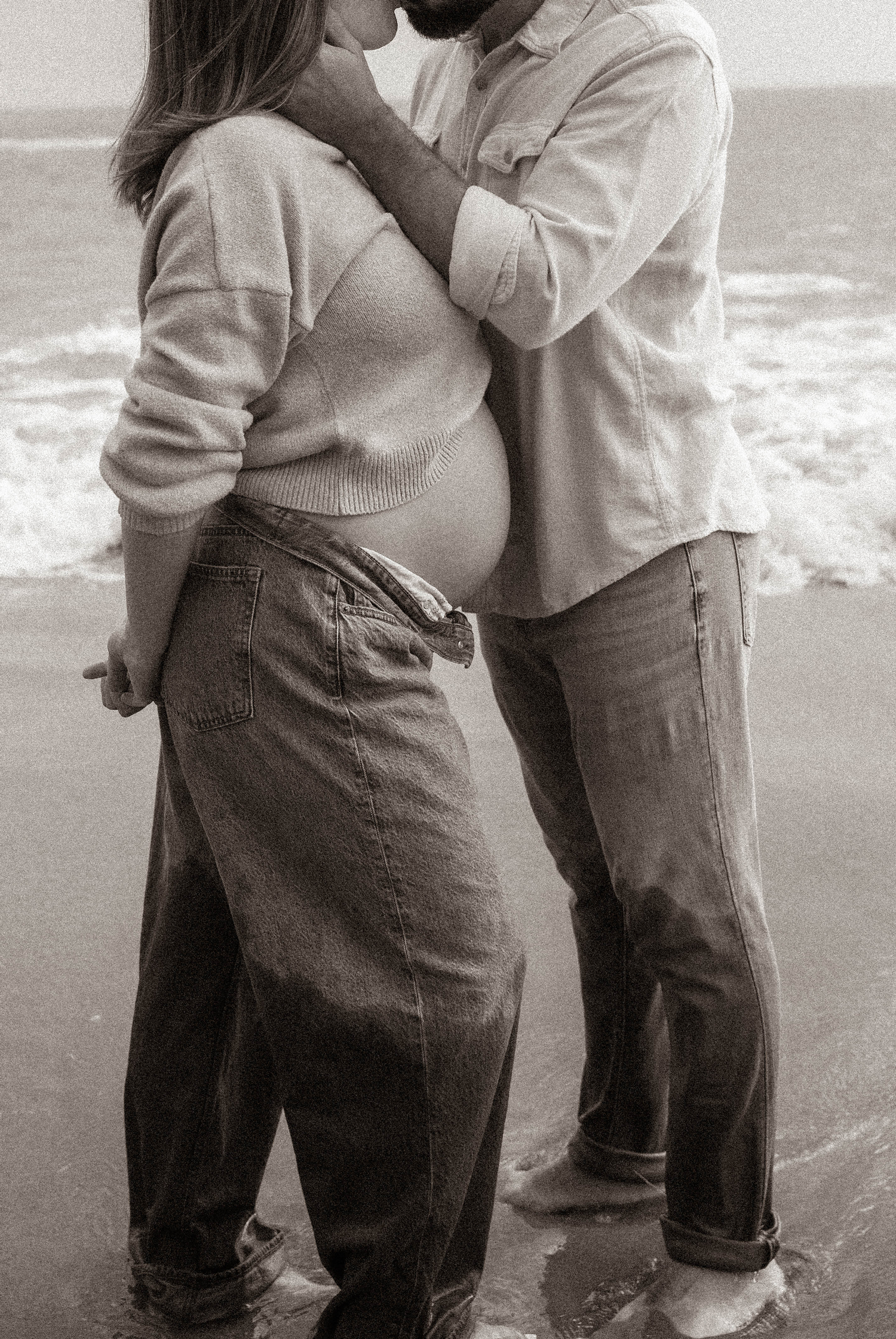 Black and white maternity photo of couple kissing on beach, man cradling pregnant woman's bare belly.