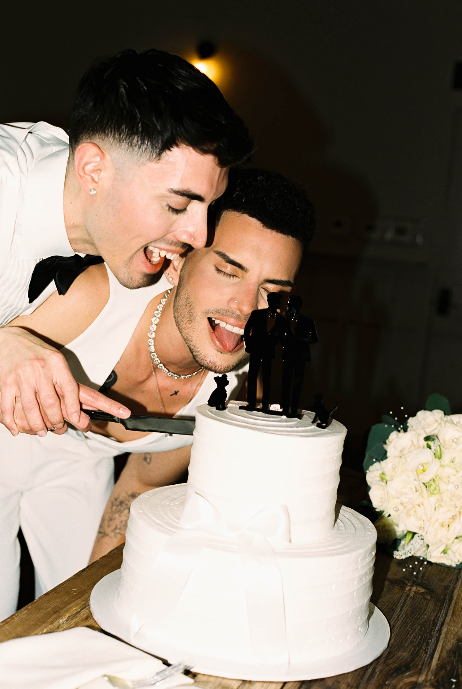 Two grooms laughing while cutting their white three-tier wedding cake topped with a same-sex couple figurine.