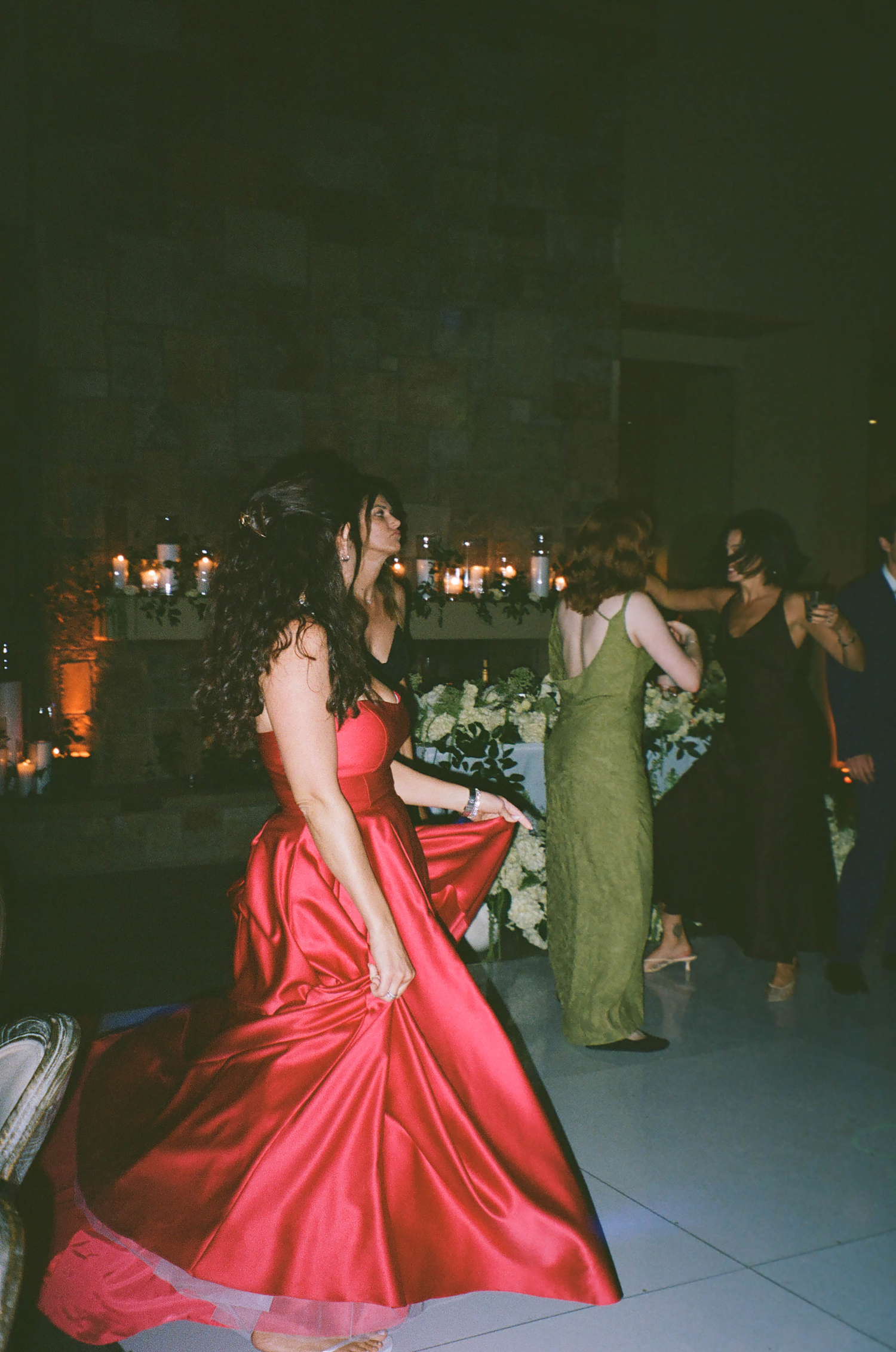 Woman in elegant red satin ball gown dancing at a formal evening event with candles and guests in background.