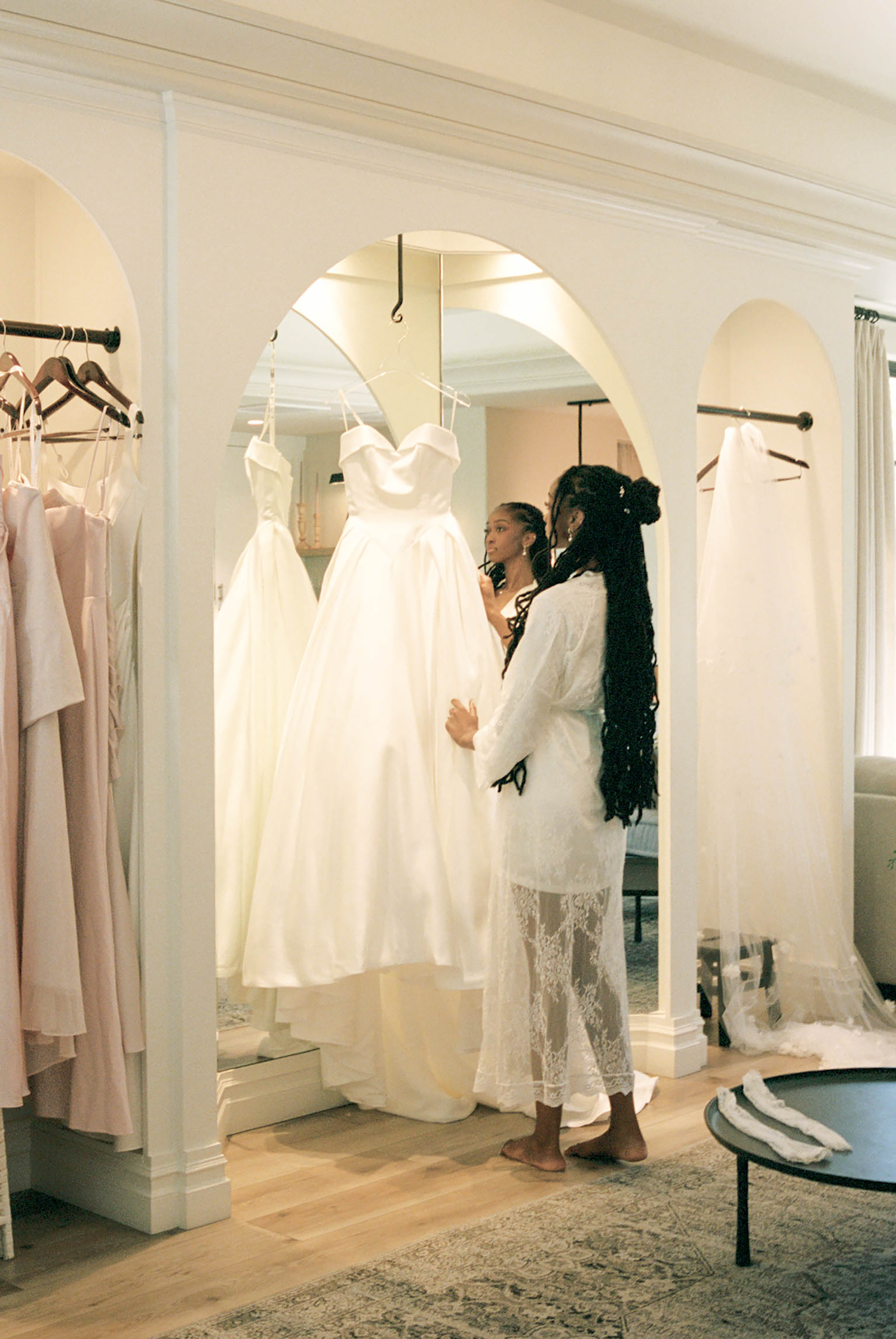 Bride trying on a white wedding gown in an elegant bridal boutique with arched mirrors and pink dresses.