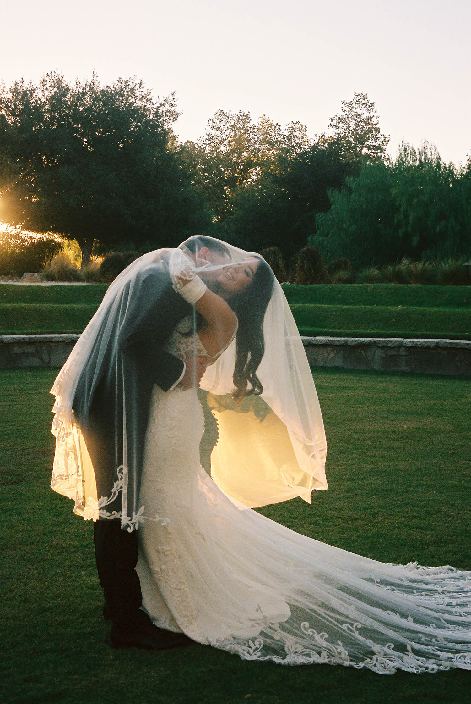 Bride and groom share a romantic kiss under a flowing lace veil at sunset in a lush garden wedding.