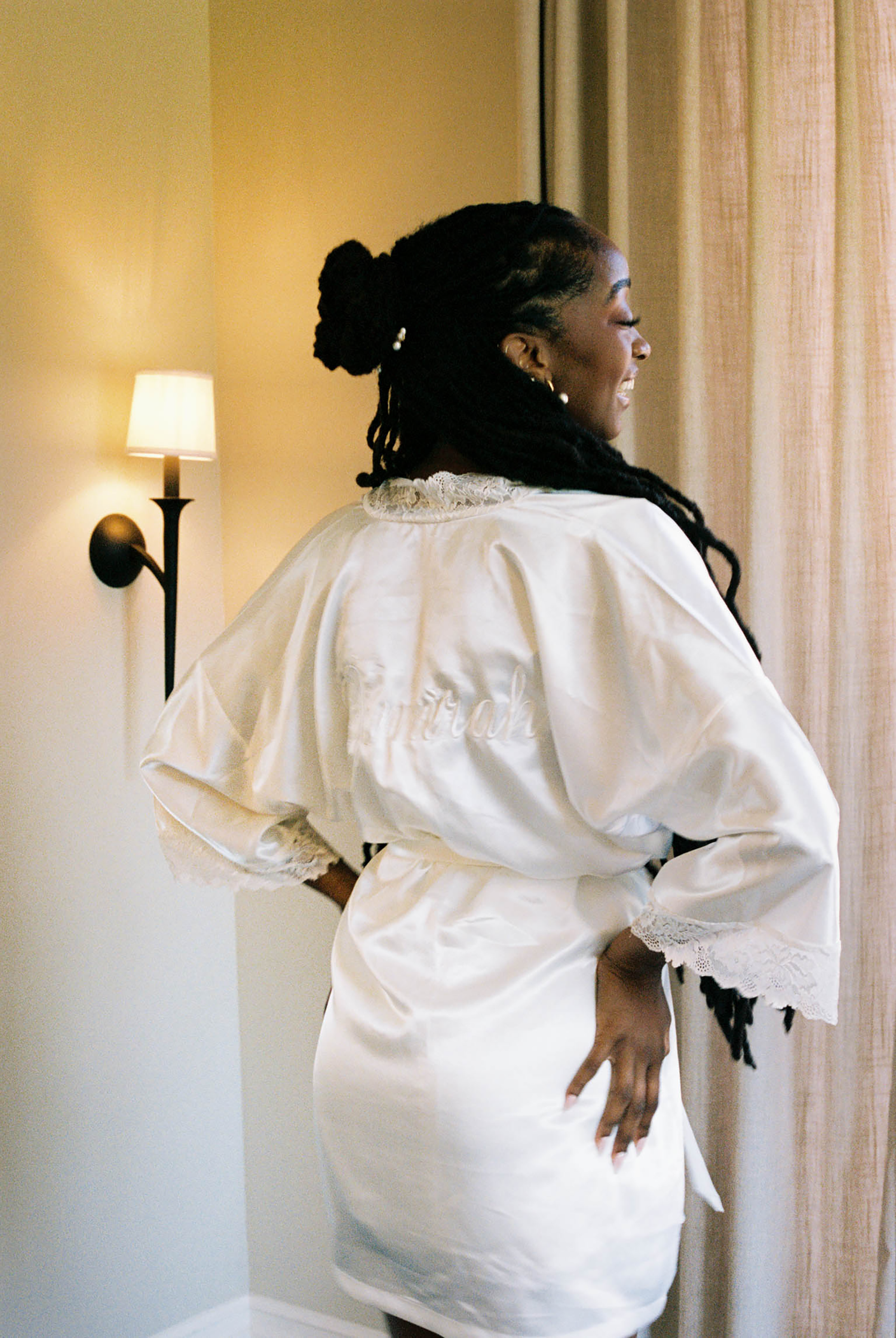 A bride in a white satin robe with lace trim stands by a window in warm light, her hair elegantly styled.