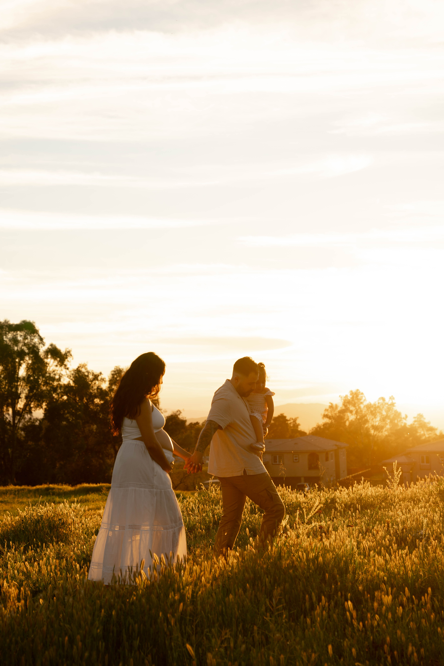 Couple holding hands in golden sunset field, woman in white dress, romantic maternity photo session.