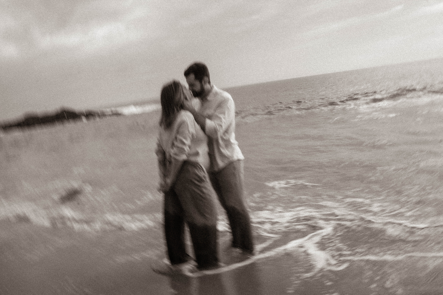Couple kissing in ocean waves on beach, romantic sepia-toned maternity photo session.