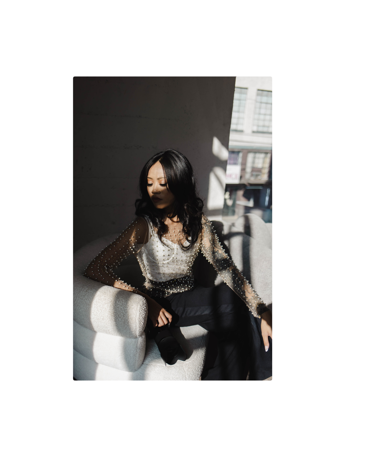 Woman in metallic mesh top and black pants sits bathed in dramatic sunlight near window.