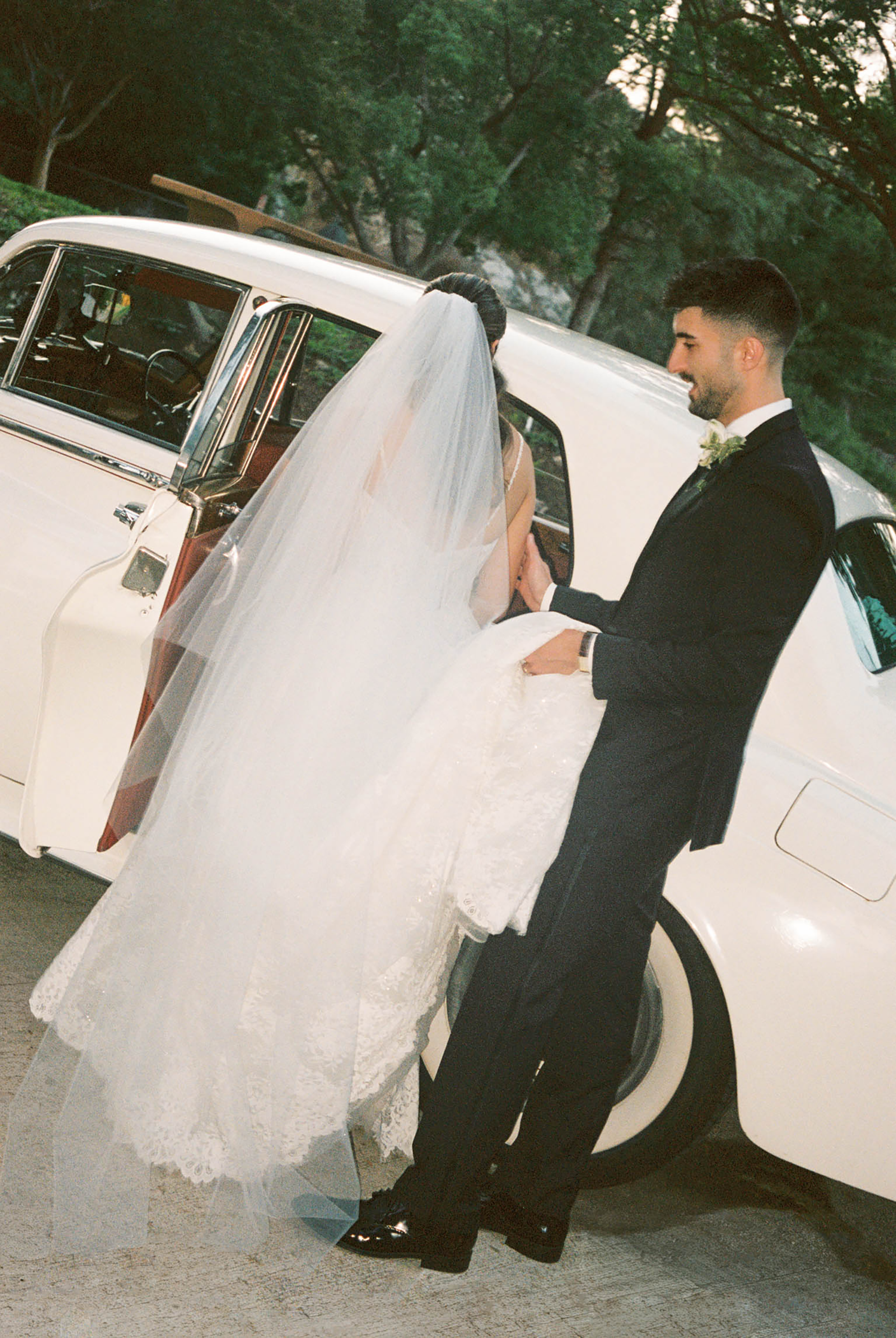 Groom helping bride into vintage white classic car, bride's veil flowing as she enters the vehicle.