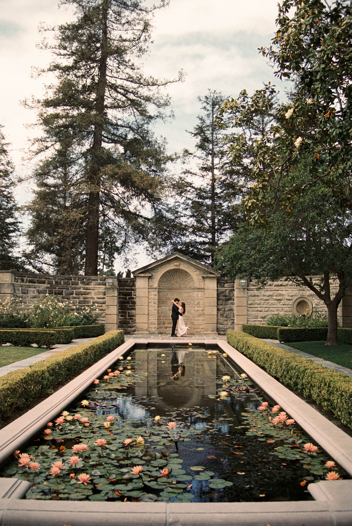 Couple embracing by a lily pond in a formal garden with stone walls and tall trees at a wedding venue.