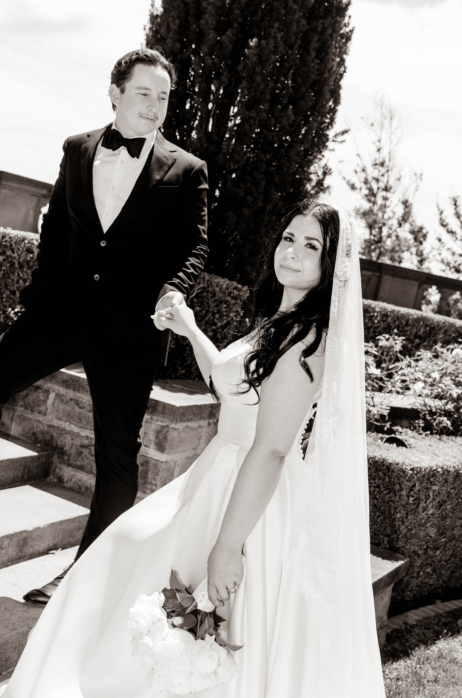 Black and white photo of a bride in white gown holding bouquet and groom in tuxedo holding hands outdoors.