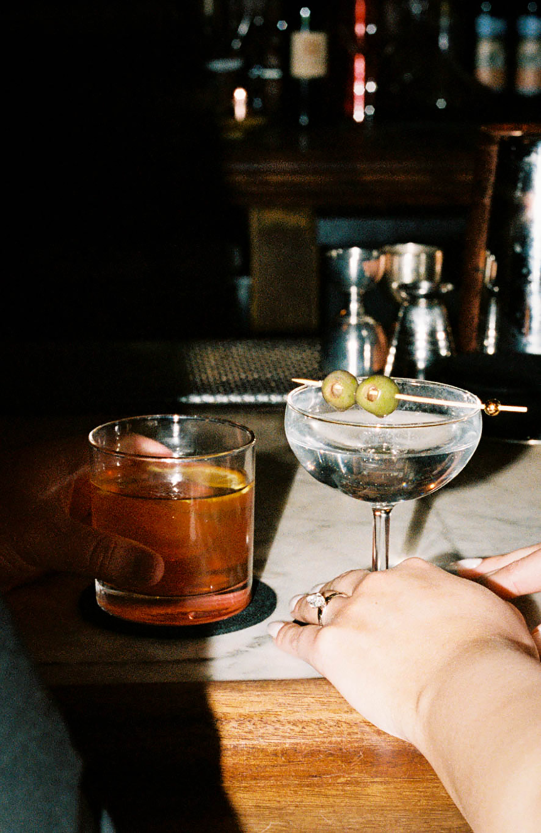 Two cocktails on a bar, a whiskey old fashioned and a dirty martini garnished with green olives on a pick.