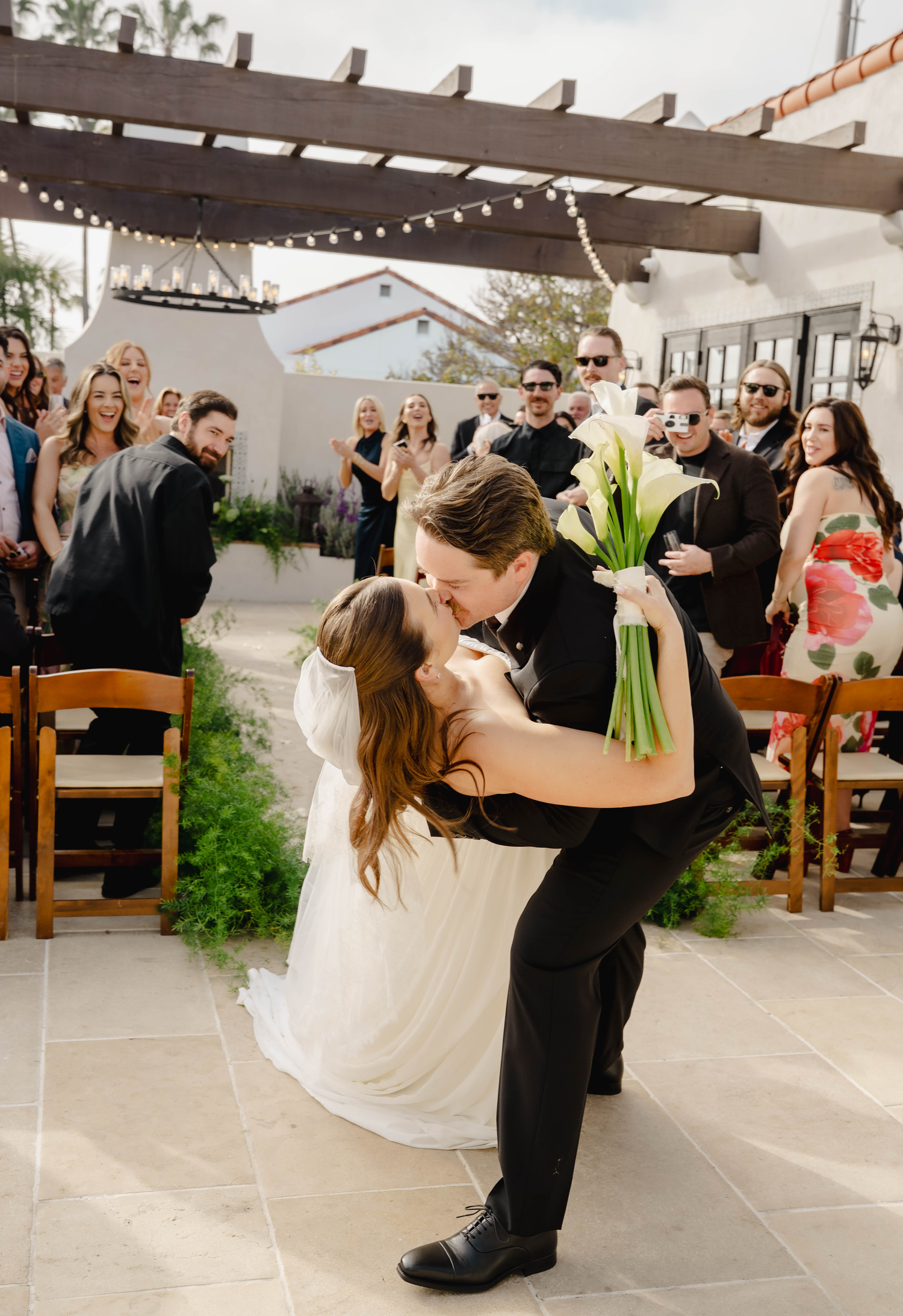 Groom dips bride for romantic kiss at outdoor wedding ceremony while guests cheer under string lights.