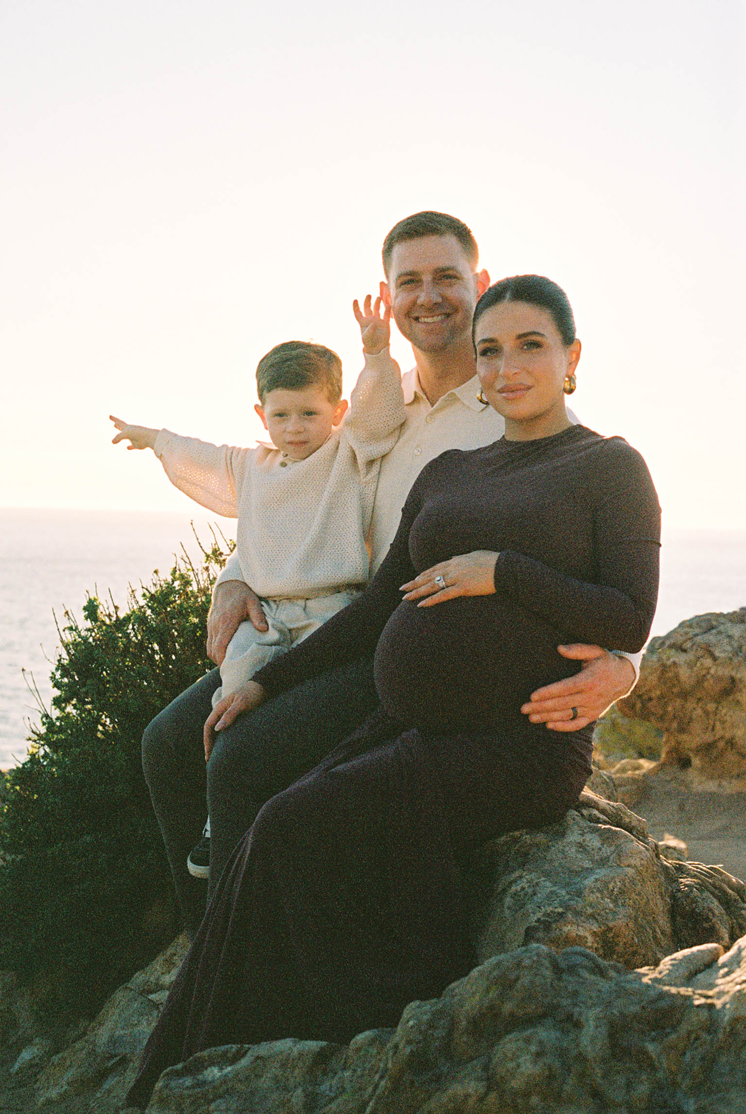 A pregnant woman in a black dress sits on rocks with her husband and young son against a sunset ocean backdrop.