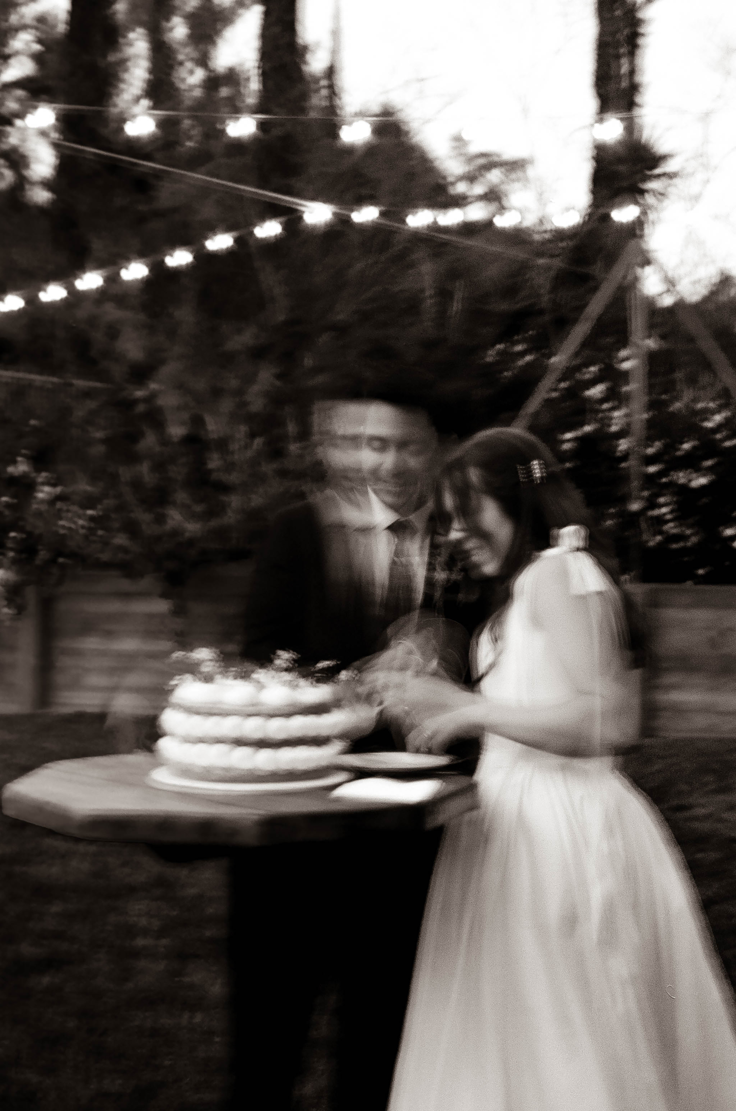 Blurry black and white photo of a bride and groom cutting their wedding cake outdoors under string lights.
