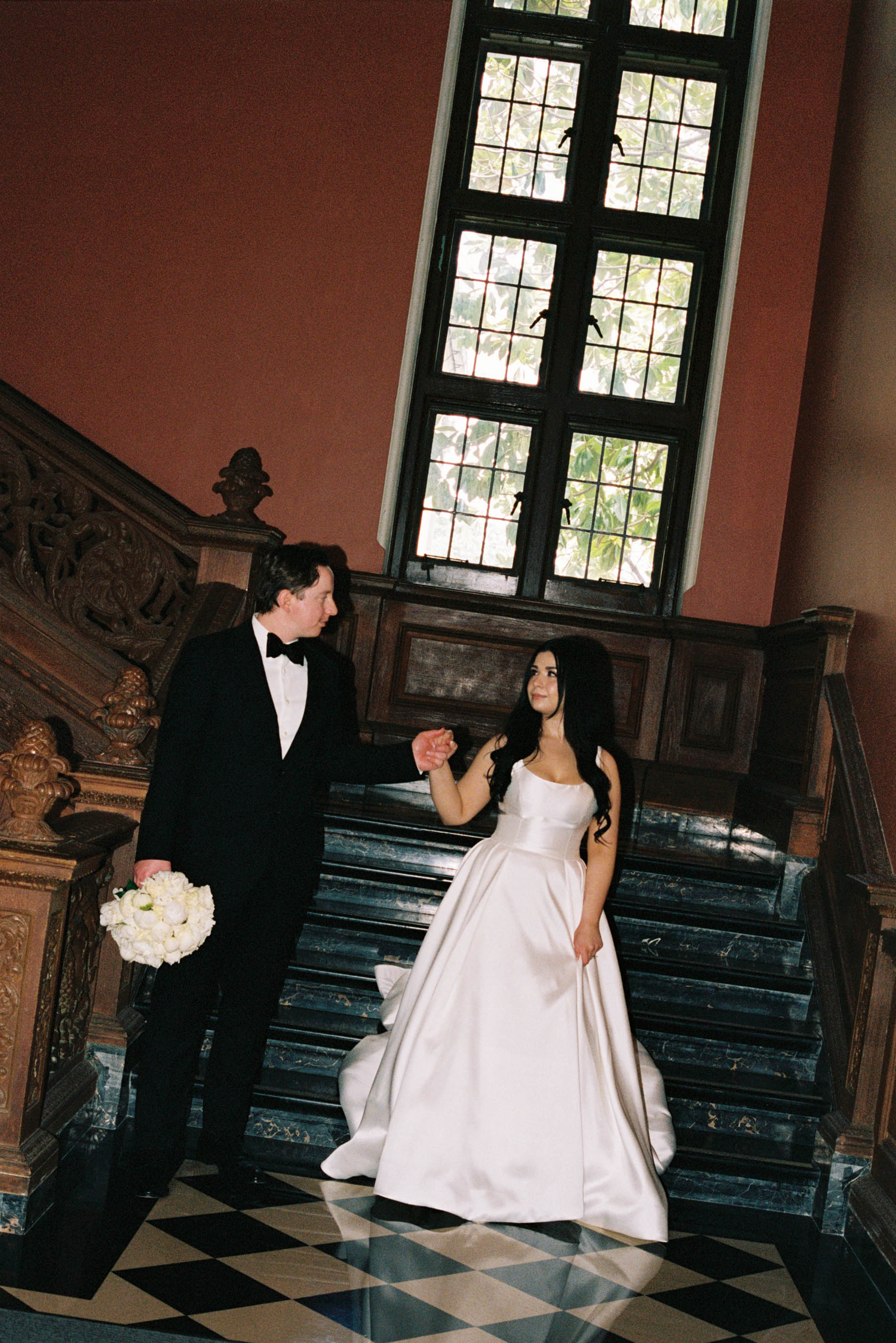 Bride and groom holding hands on grand marble staircase, she in white ball gown, he in black tuxedo, elegant venue.