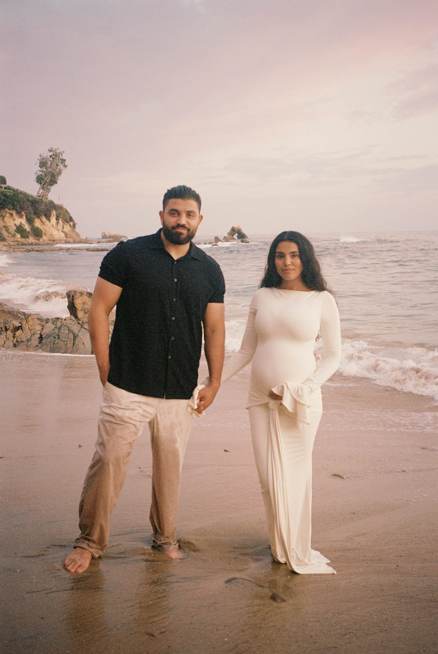 A couple holds hands on a sandy beach at sunset, woman in white maternity gown showing her bump.