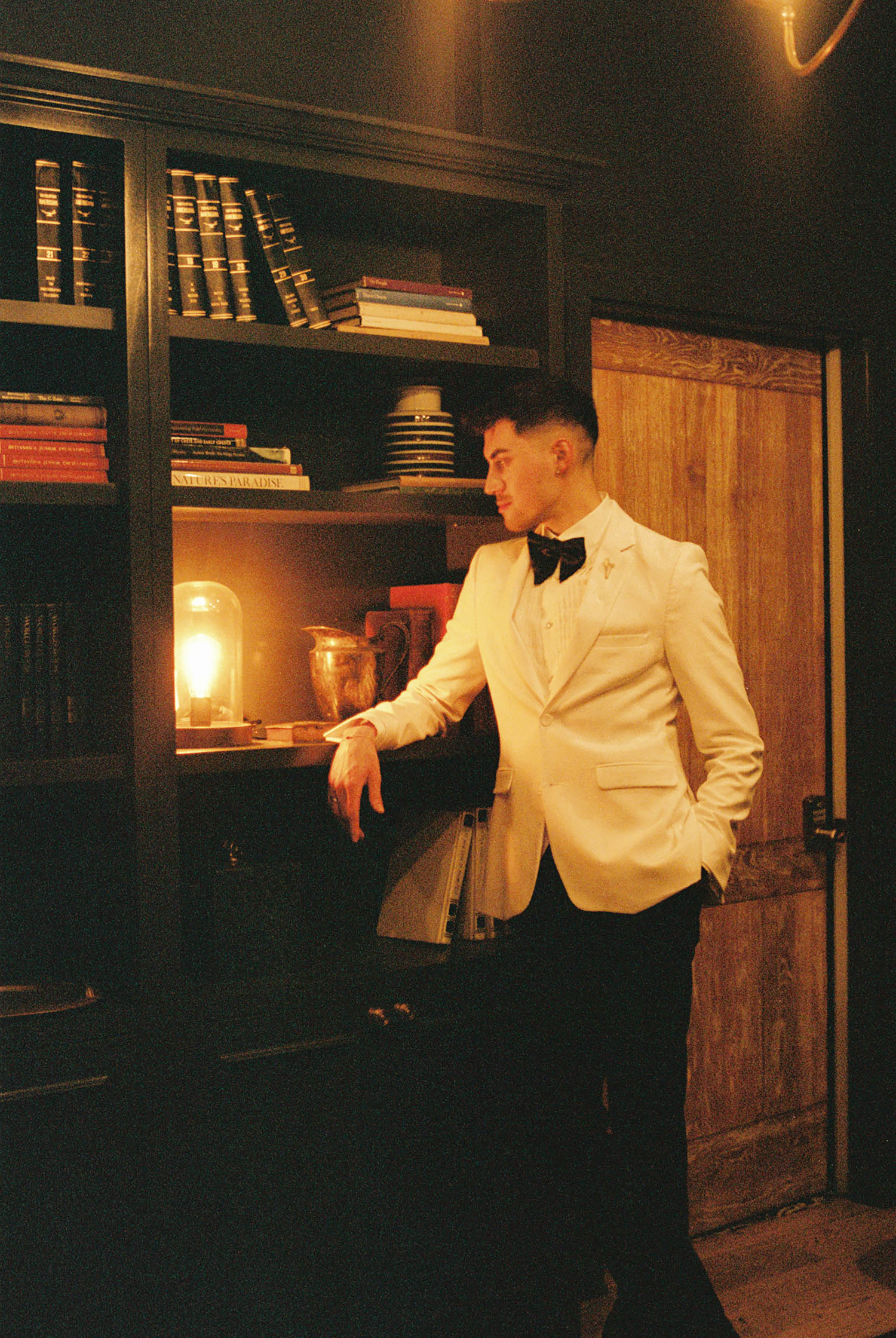 Man in white tuxedo jacket and black bow tie poses by a dimly lit bookshelf in an elegant library.