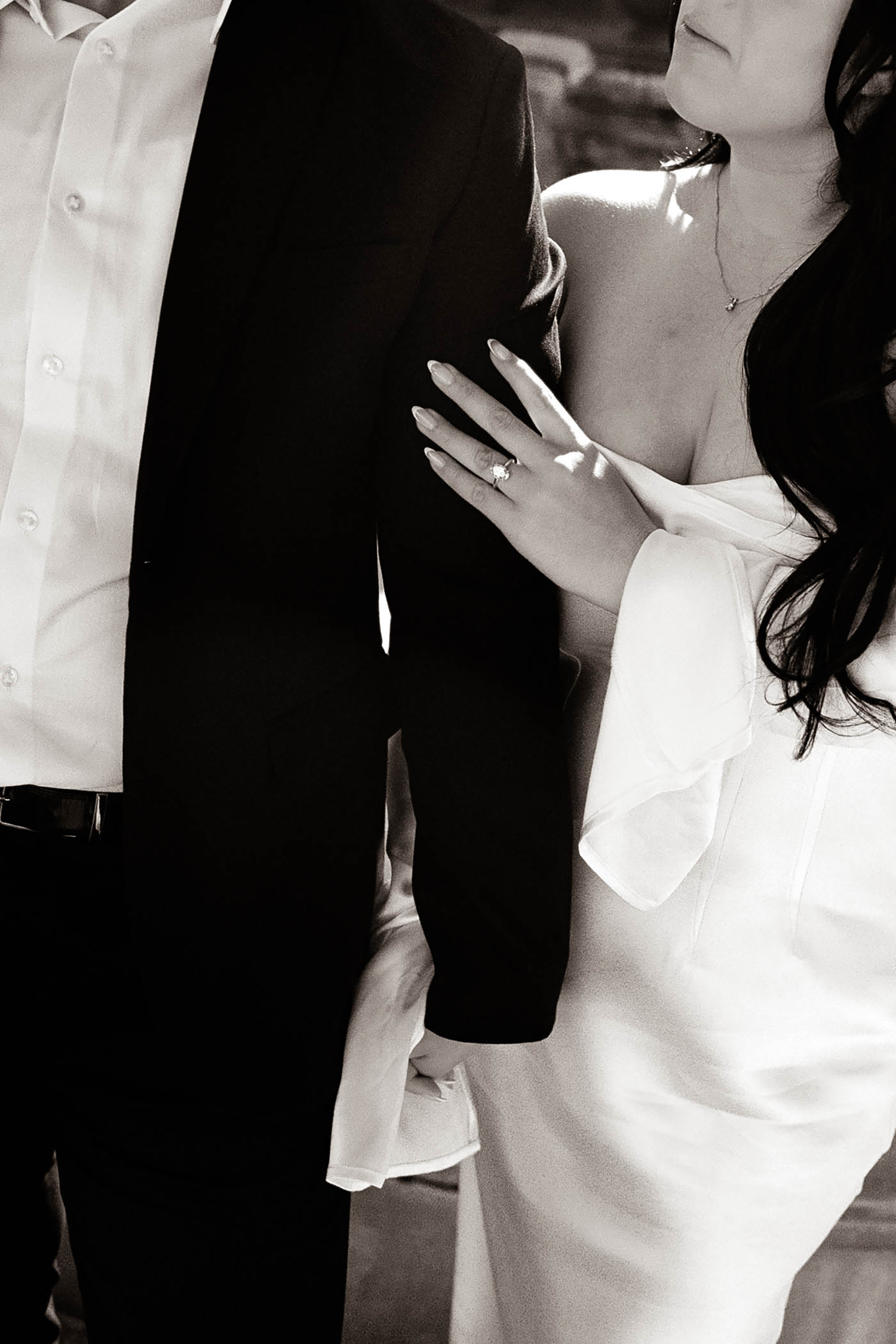 Black and white close-up of bride in white dress placing hand on grooms black suit jacket. Classic & editorial.