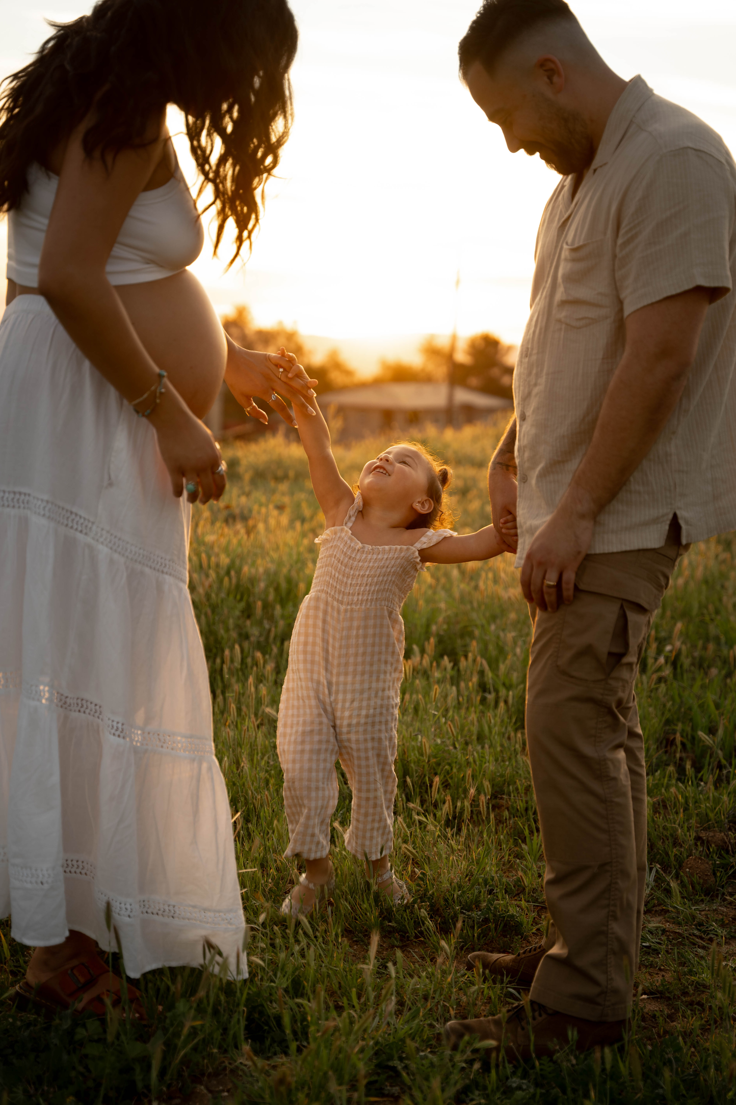 Pregnant woman in white dress and man hold toddler girl's hands in golden sunset field.