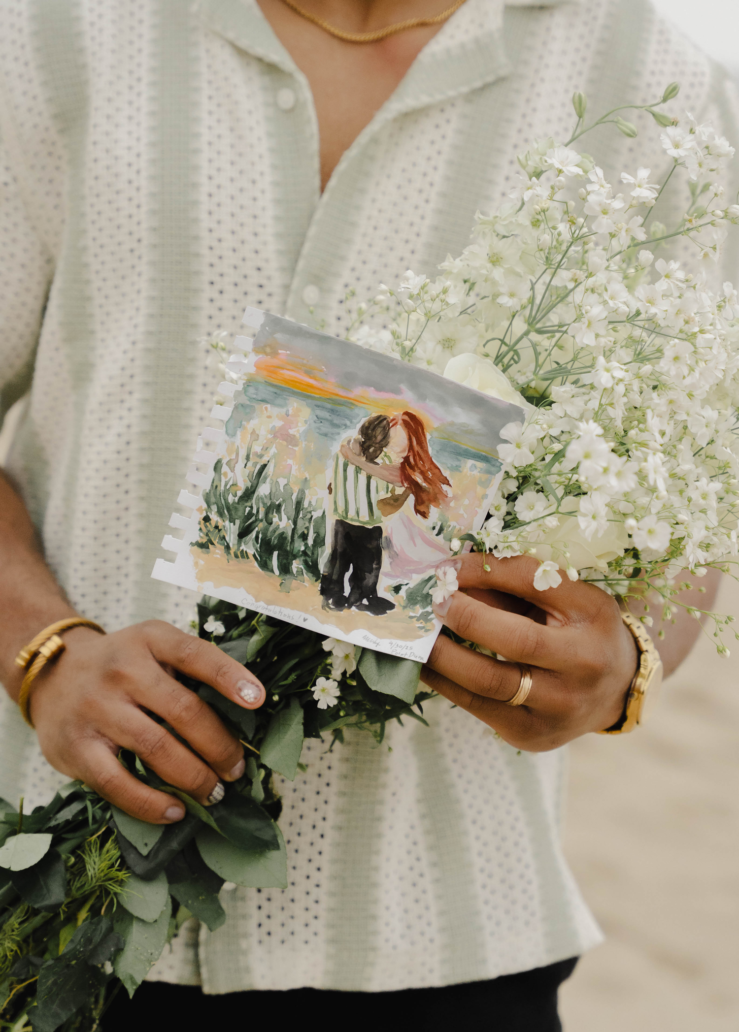 Person holding a romantic couple painting and white wildflower bouquet, wearing a gold ring and bracelet.