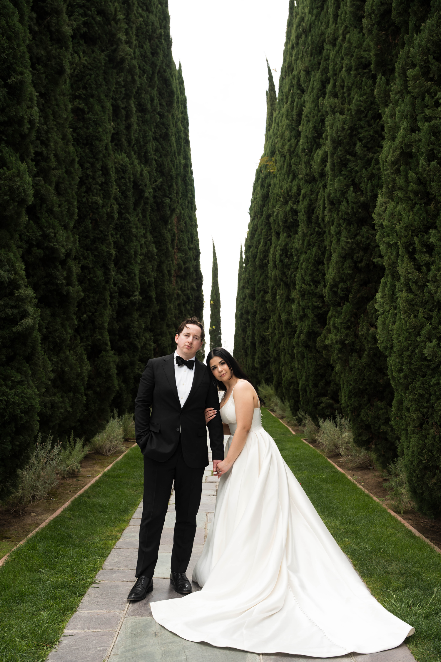 Bride and groom pose elegantly on a garden path lined with tall cypress trees in a romantic outdoor wedding portrait.