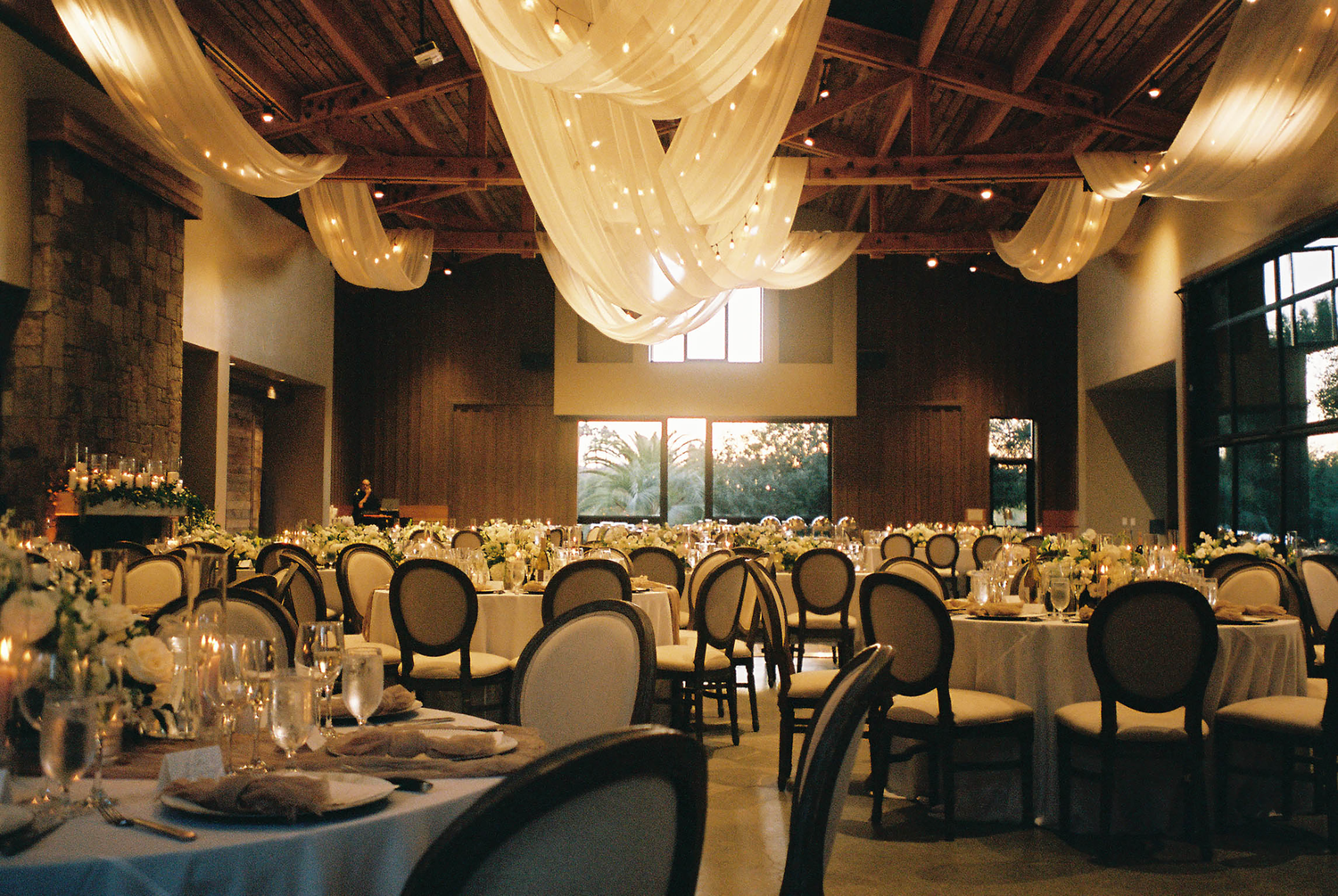 Elegant wedding reception venue with draped white fabric ceiling, round tables, gold chairs, and floral centerpieces in warm lighting.
