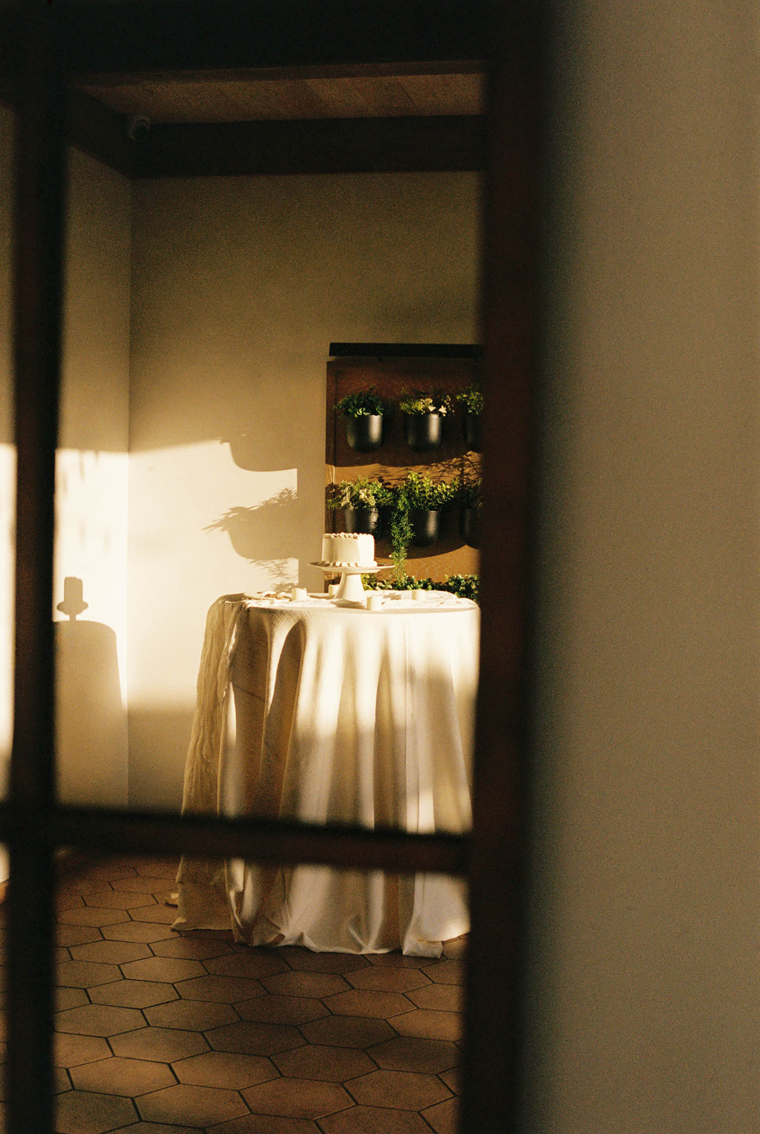 Elegant wedding cake table with white linen and greenery wall backdrop in warm golden light.