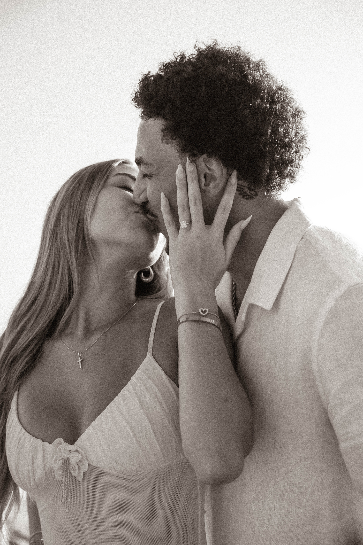 Black and white photo of couple kissing, woman showing engagement ring on beach.