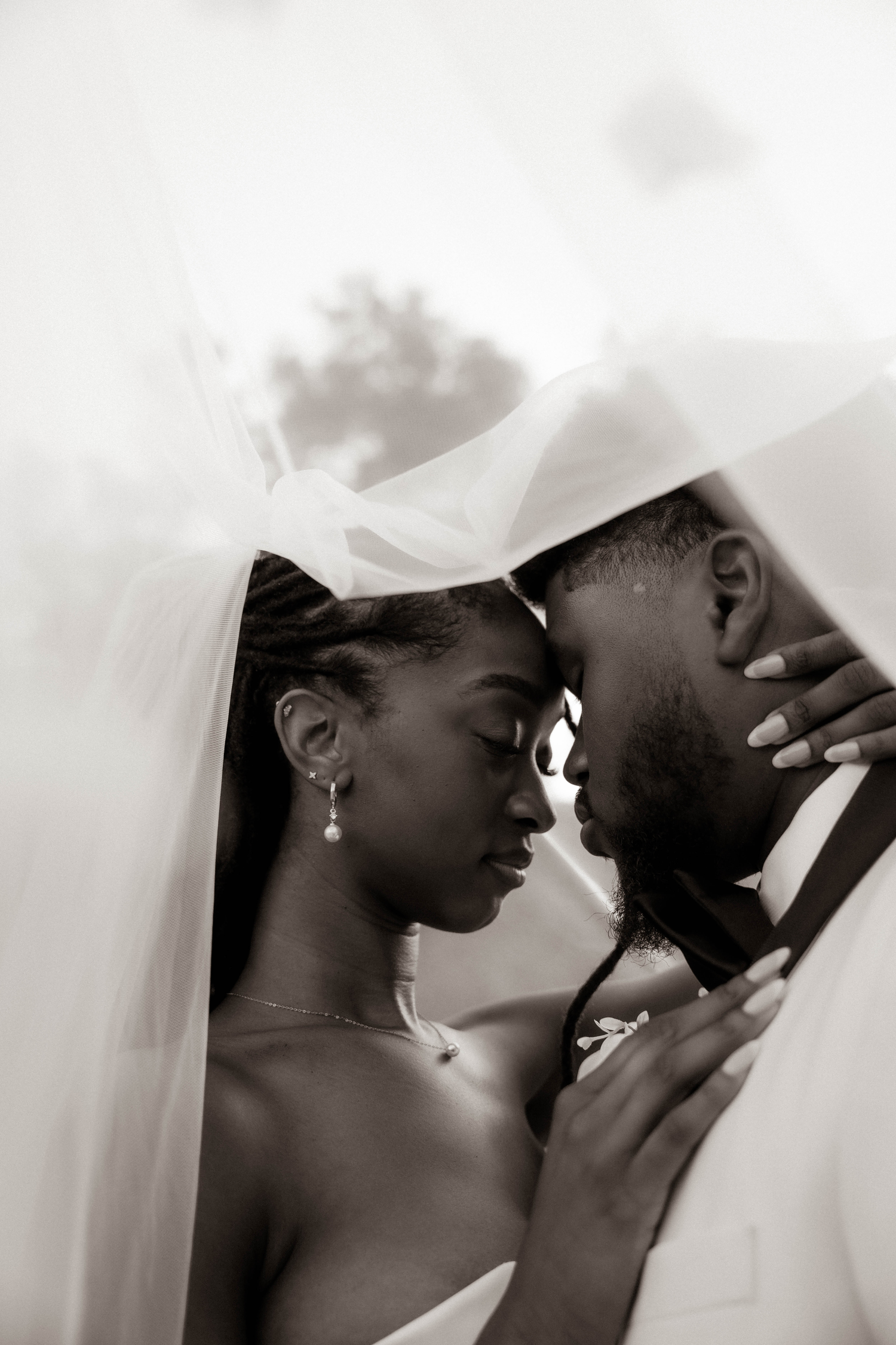 Black and white wedding photo of a bride and groom touching foreheads tenderly under a flowing veil.