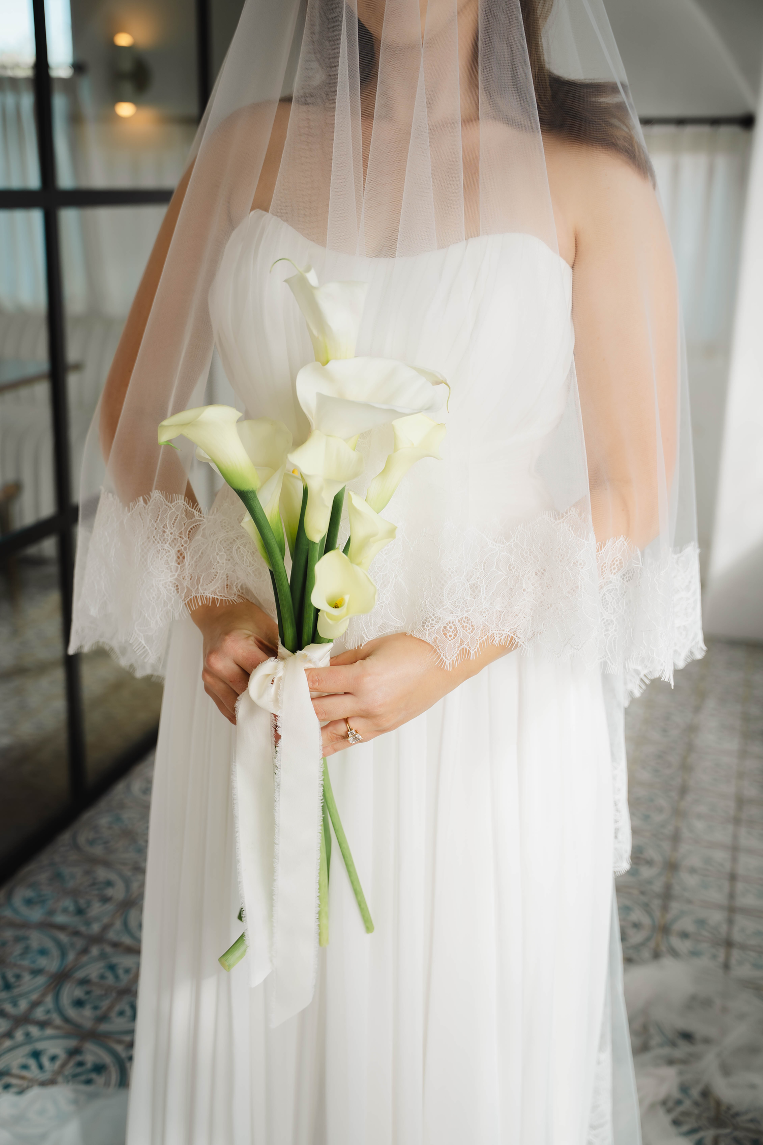 Bride in strapless white gown holding elegant white calla lily bouquet with lace-trimmed veil.