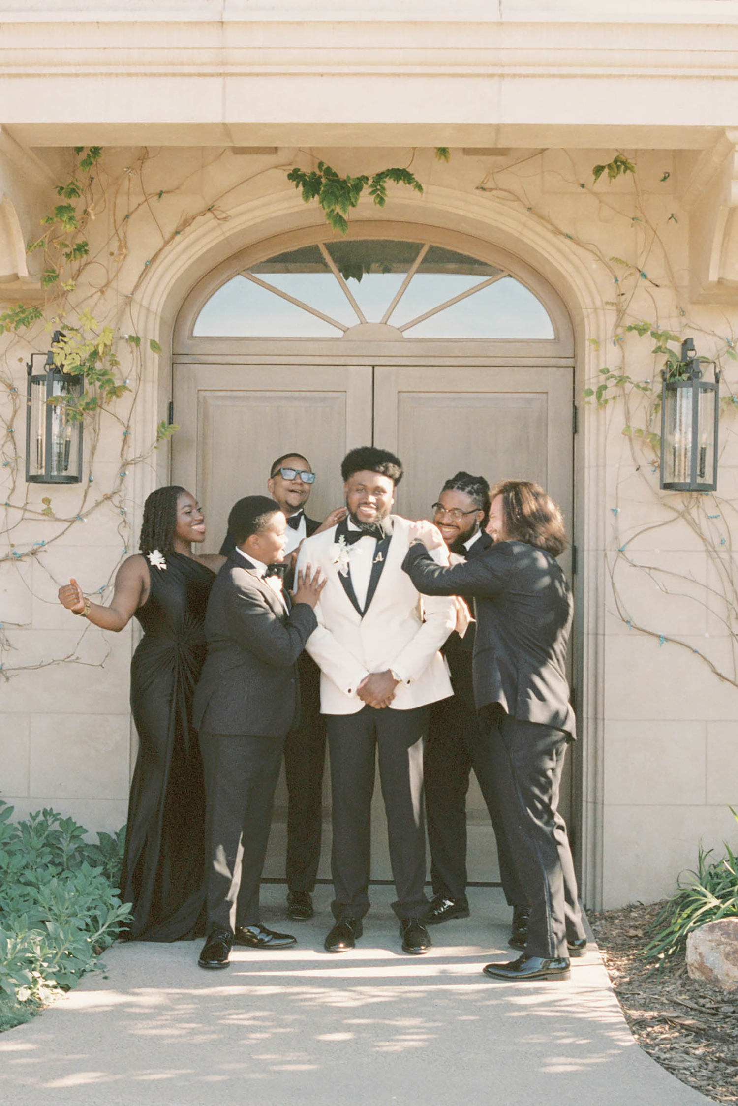 Groom in white tuxedo jacket surrounded by smiling wedding party in black attire outside elegant venue doors.