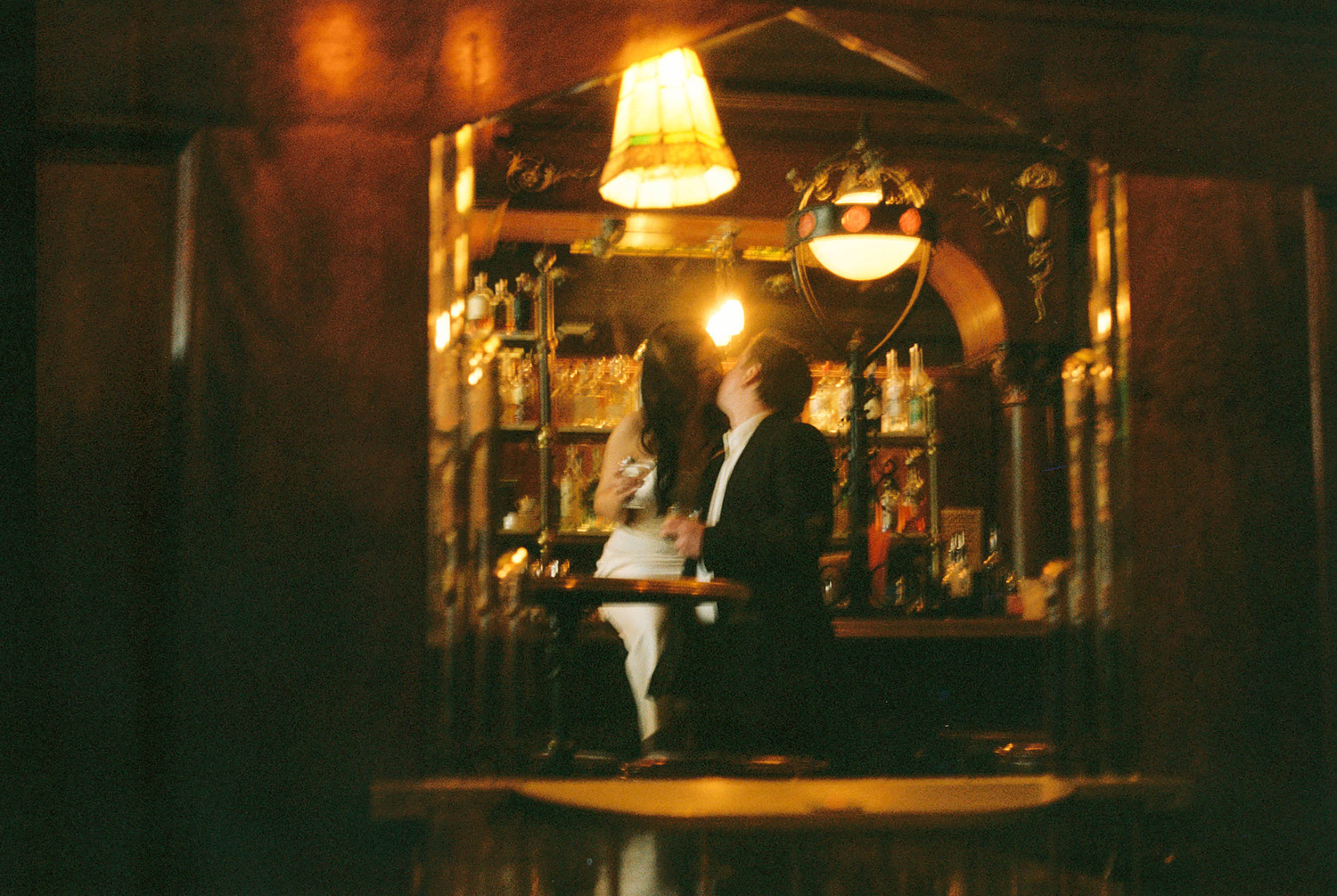 A bartender stands behind a warmly lit bar, illuminated by a hanging lamp in a cozy, dimly lit establishment.