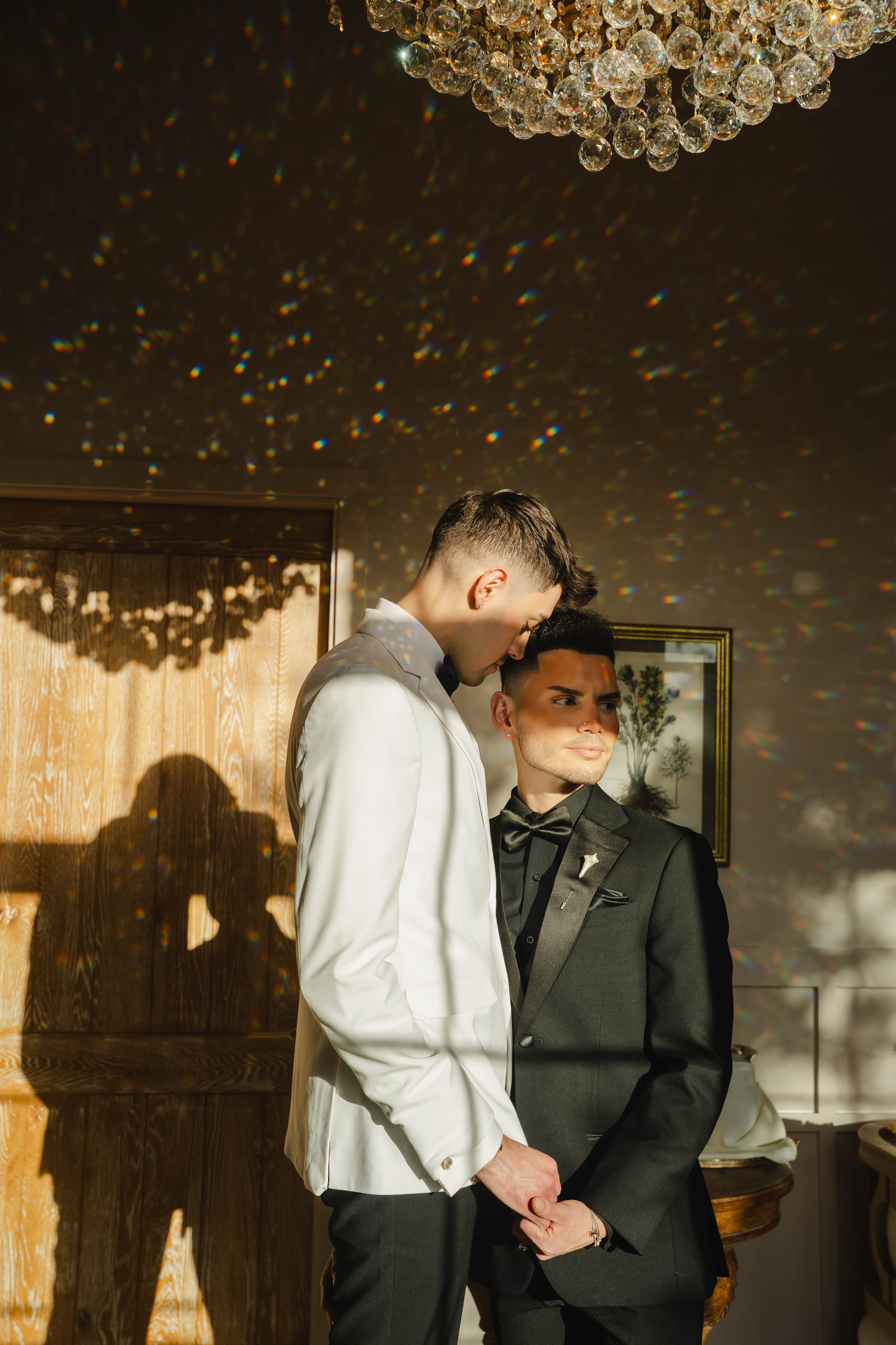 Two grooms share an intimate moment indoors, one in a white shirt and one in a black tuxedo, under a crystal chandelier.