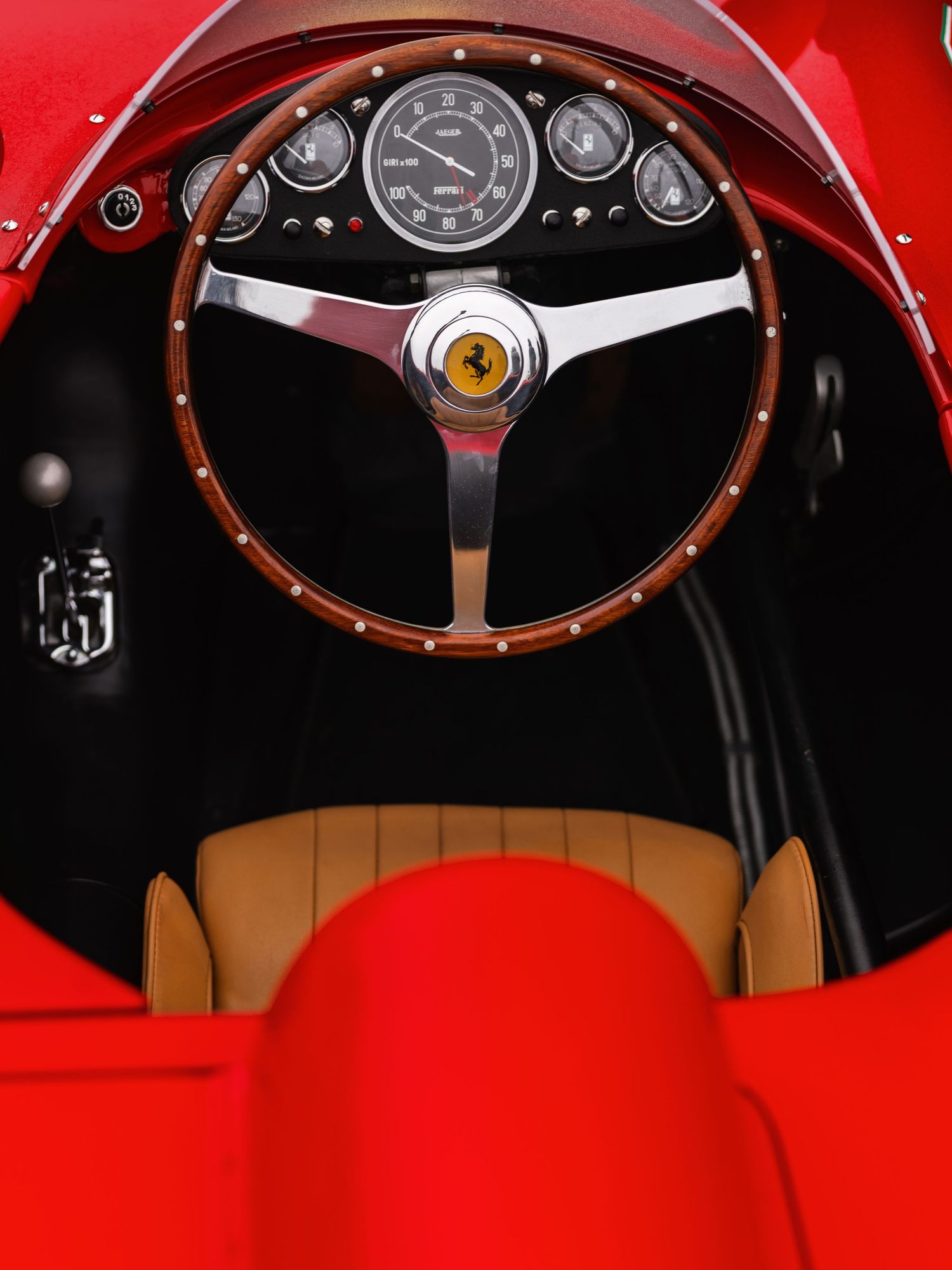 Vintage Ferrari cockpit with wooden steering wheel, prancing horse emblem, classic gauges, and tan leather seat interior.