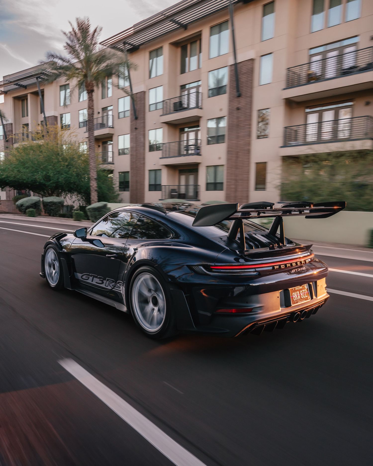 Black Porsche 911 GT3 RS driving on a city street with palm trees and modern apartment buildings in the background.