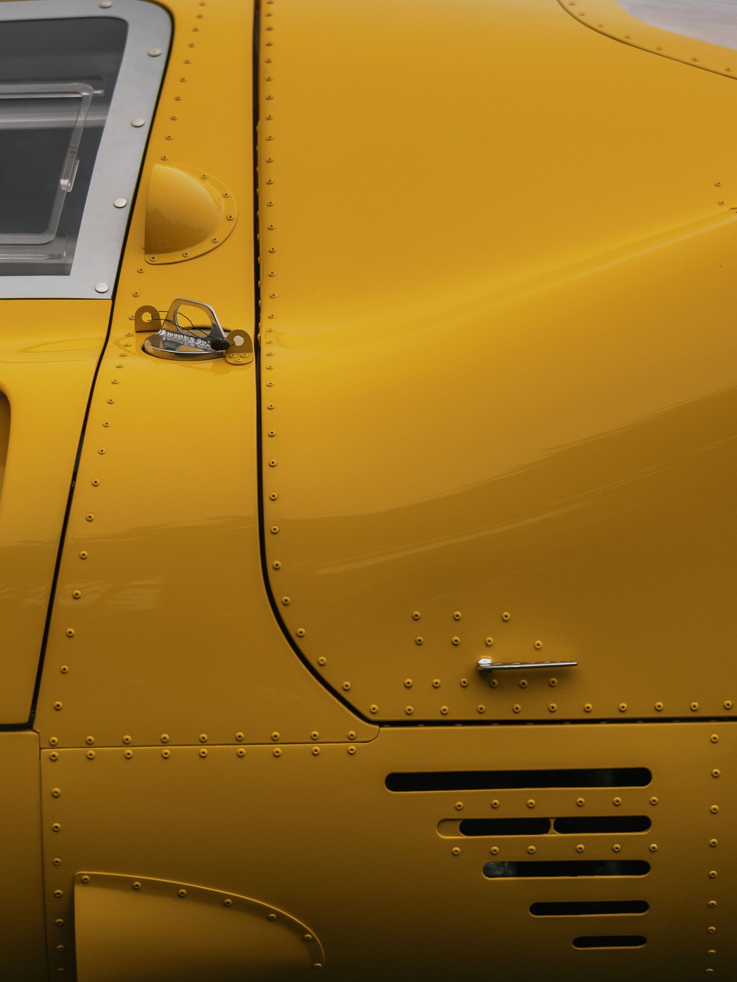 Close-up of a vintage yellow vehicle door with chrome handle, rivets, and ventilation slots on polished metal body.