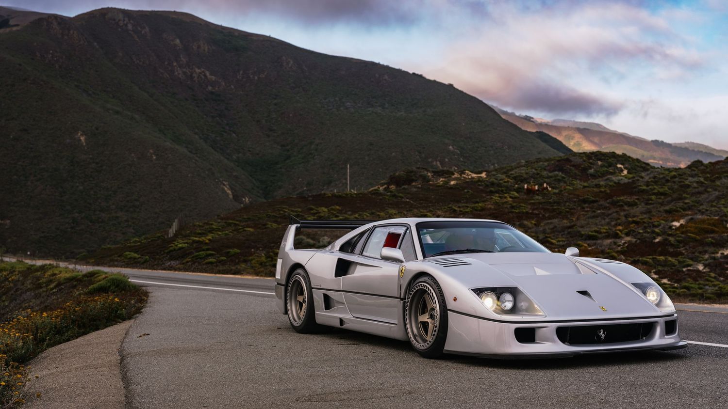 White Ferrari F40 supercar parked on a mountain road with dramatic cloudy skies and green hills in the background.