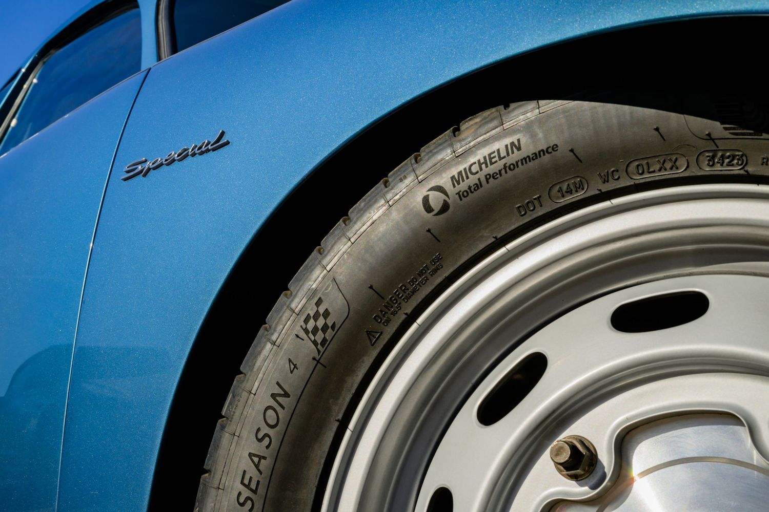 Close-up detail shot of a blue Barracuda classic car showing part of its chrome badge and white wheel rim.