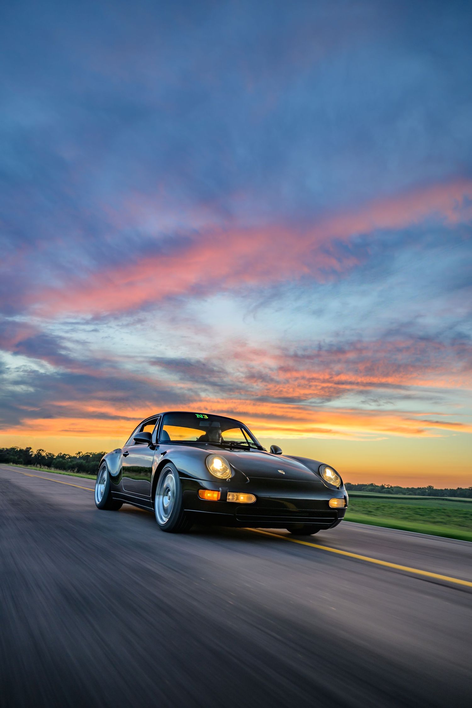 Black Porsche 911 sports car driving on road at sunset with dramatic orange and blue sky in background.