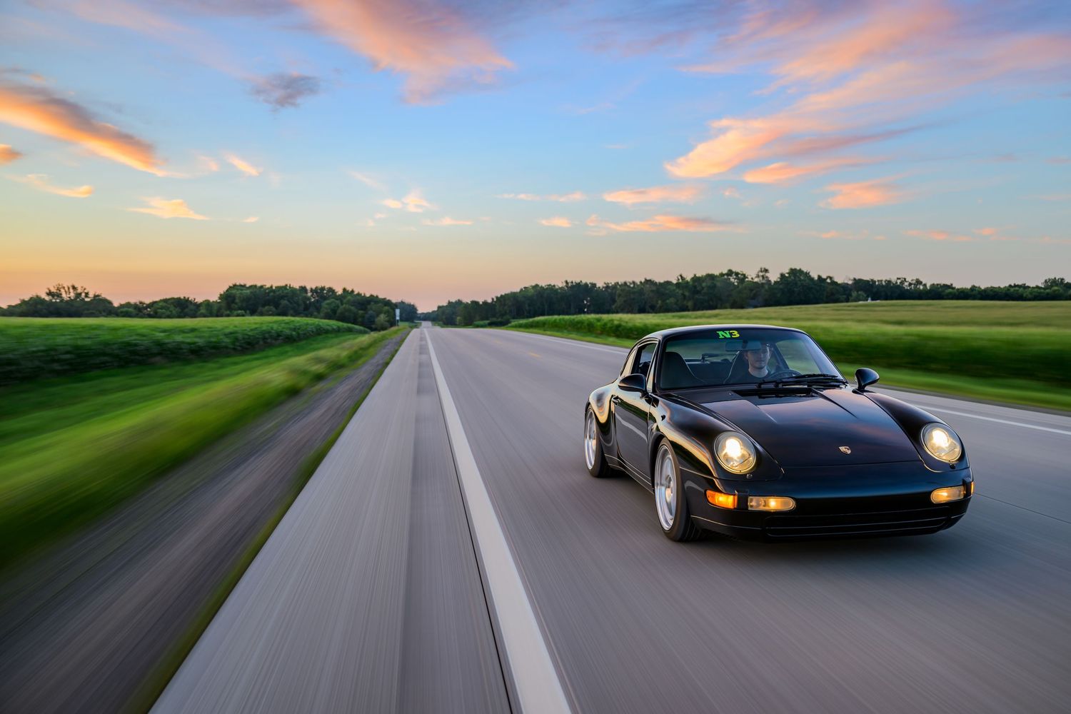 Black Porsche 911 speeding down empty highway at sunset with colorful sky and green fields in background.