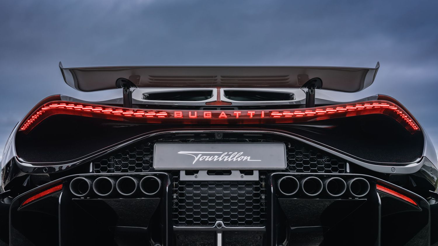 Rear view of a Bugatti Tourbillon supercar featuring red LED taillights, large rear wing, and quad exhaust pipes against a cloudy sky.