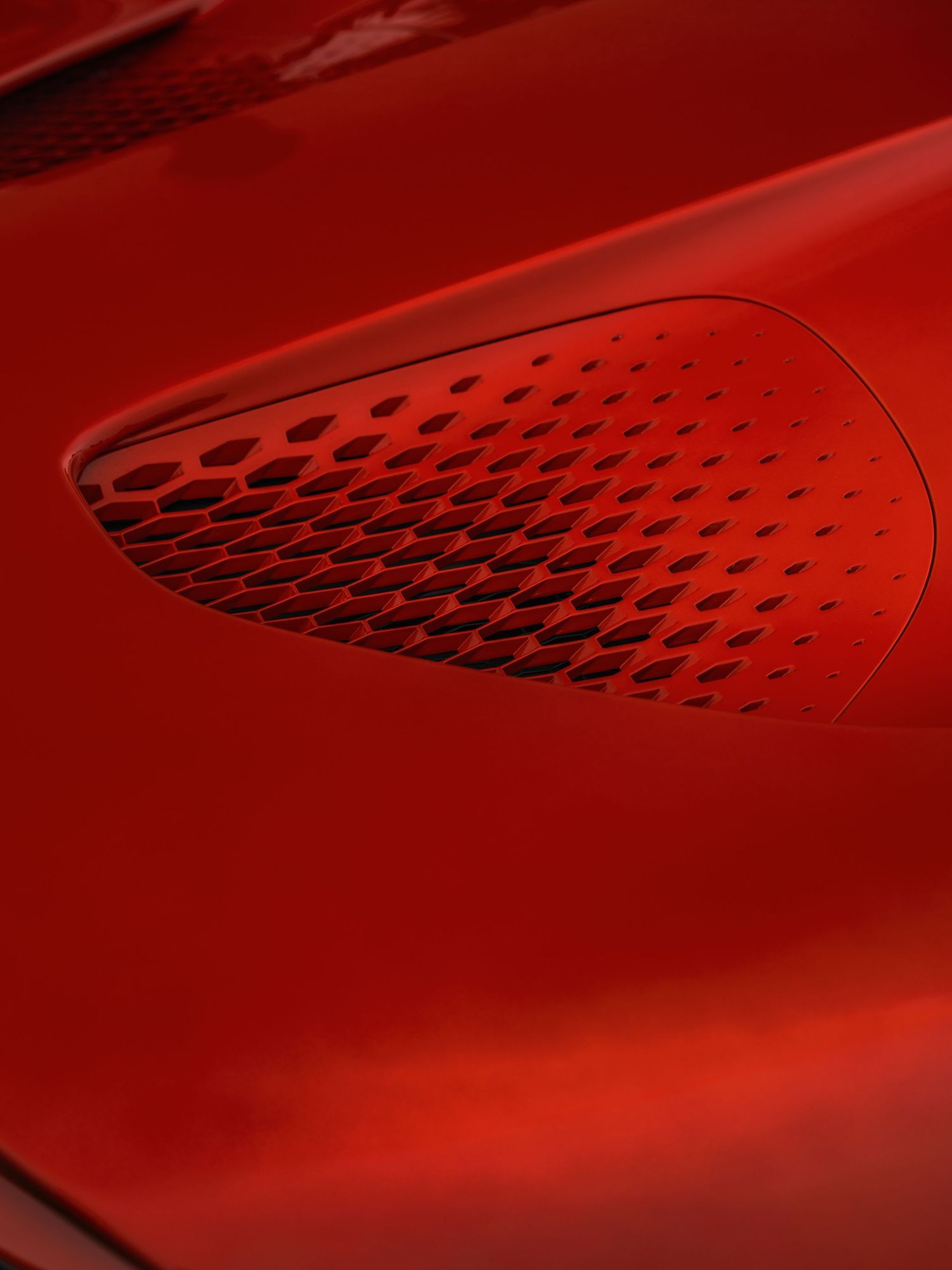Close-up of a red sports car hood vent with geometric honeycomb perforations on a glossy red surface.