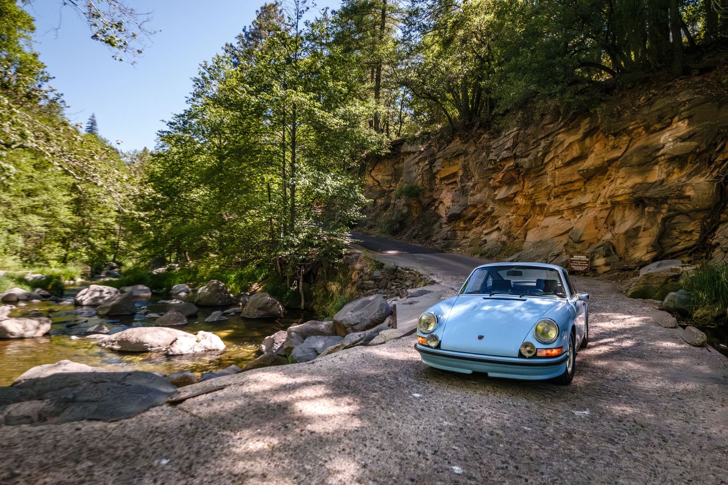 Vintage light blue Porsche 911 parked on rocky terrain beside a stream surrounded by lush green trees and sandstone cliffs.