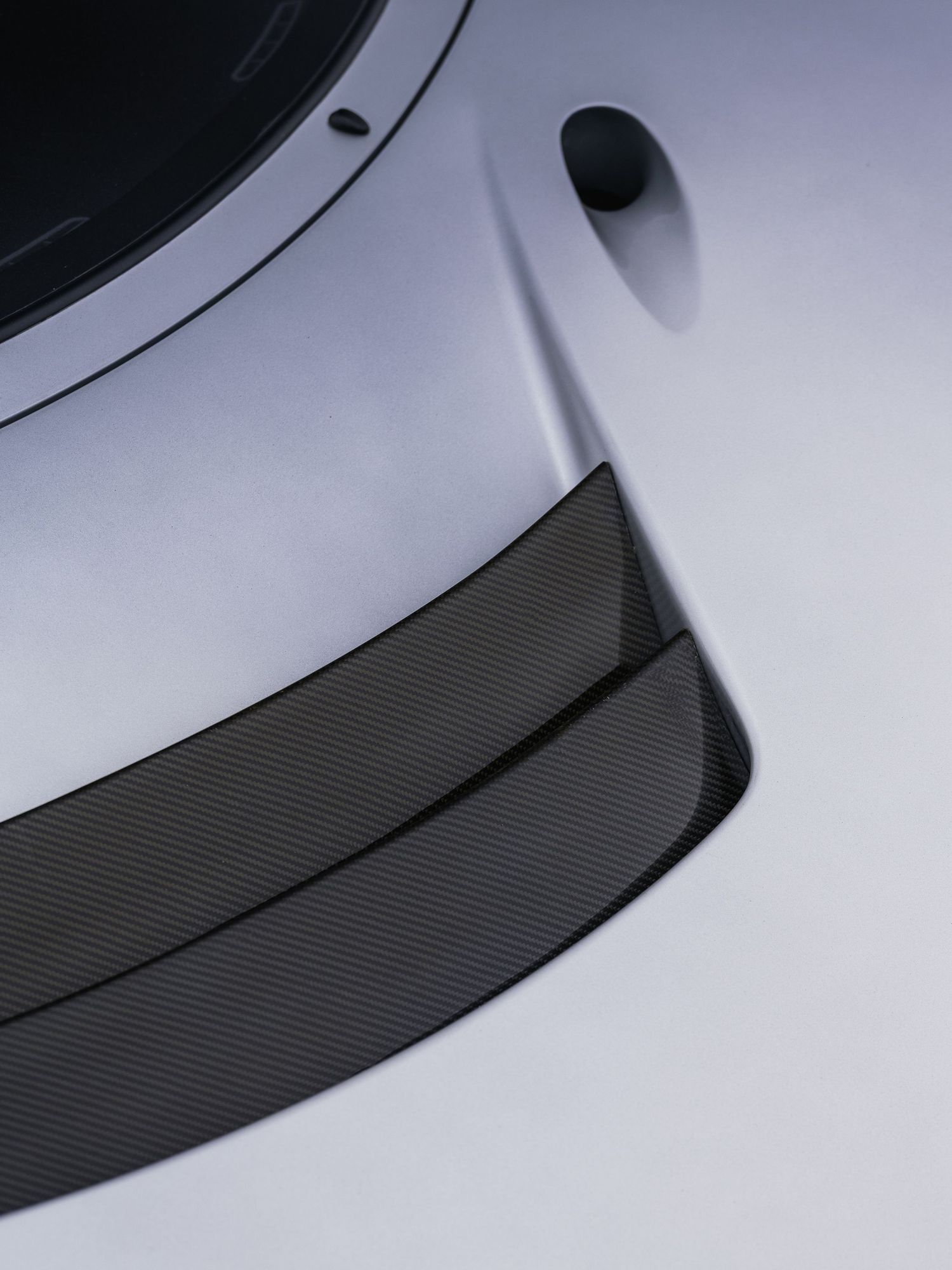 Close-up of a sleek silver car hood featuring a carbon fiber front splitter with a subtle textured weave pattern.