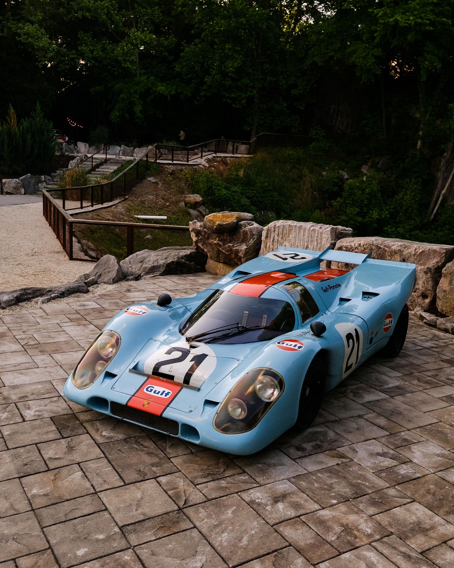 Gulf-liveried Porsche 917 race car in iconic blue and orange, number 21, parked on stone pavers surrounded by lush greenery.