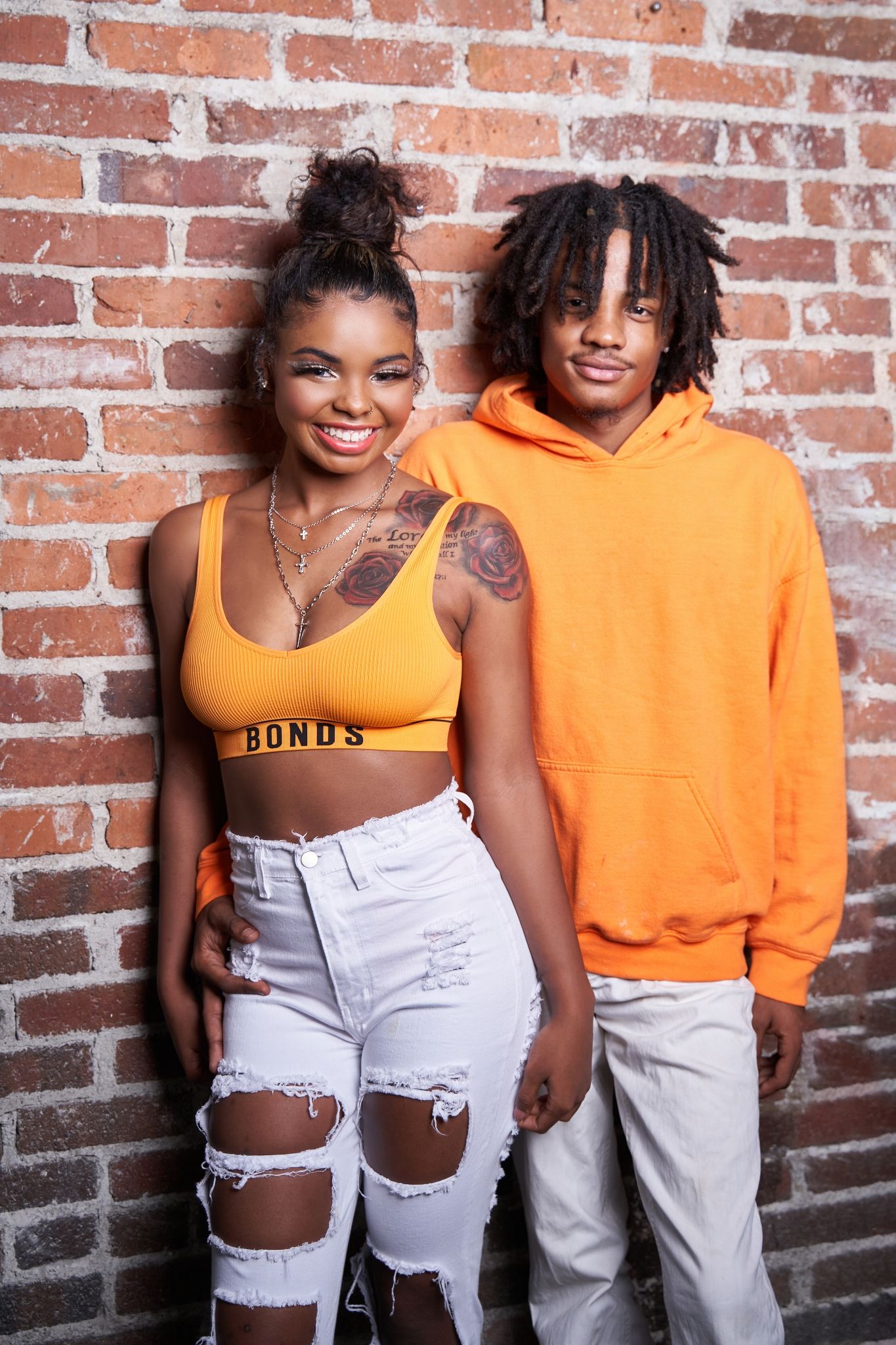Urban fashion photoshoot with bright orange hoodie and distressed white jeans against brick wall.