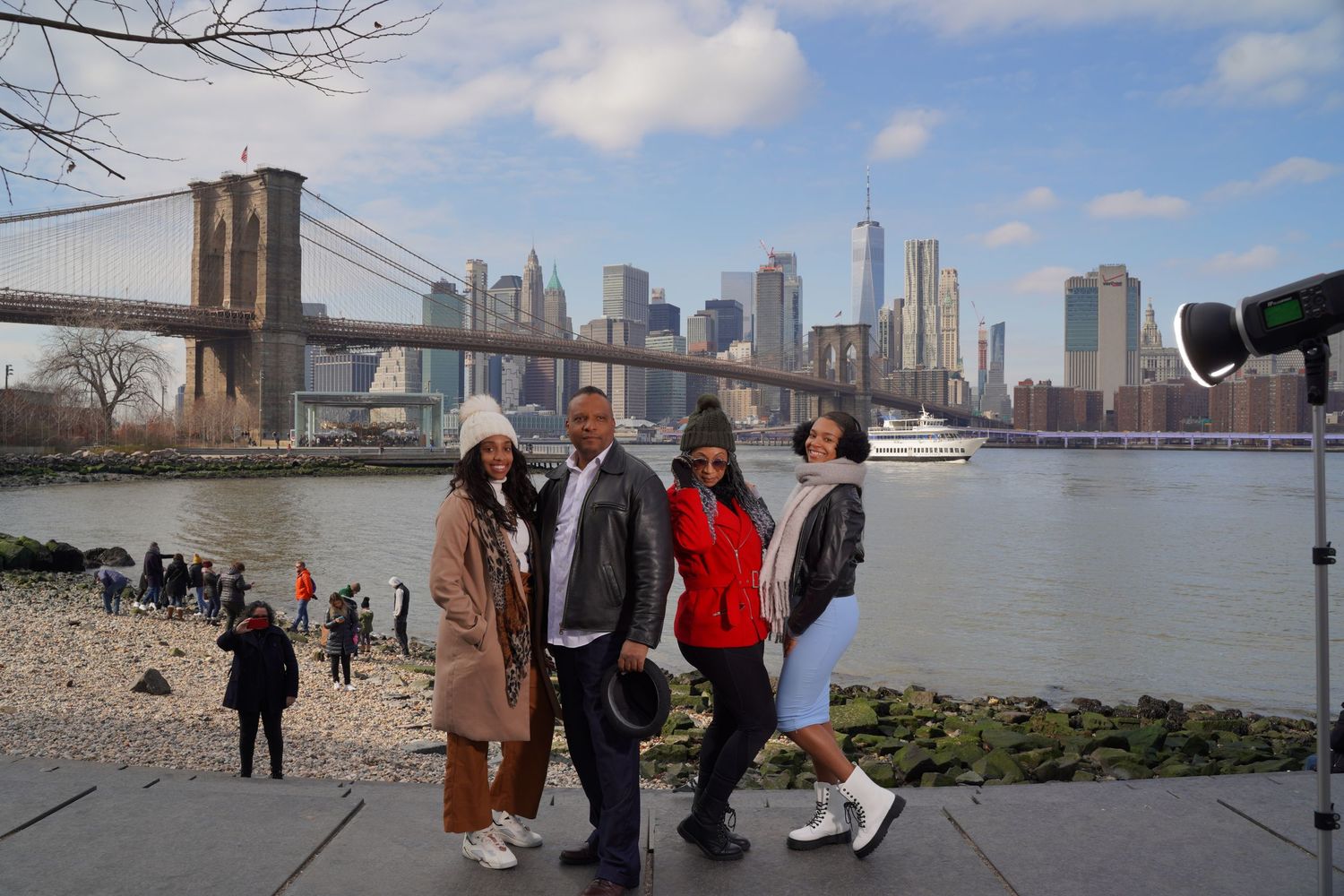 Group gathering near Brooklyn Bridge Park with Manhattan skyline visible across the river.
