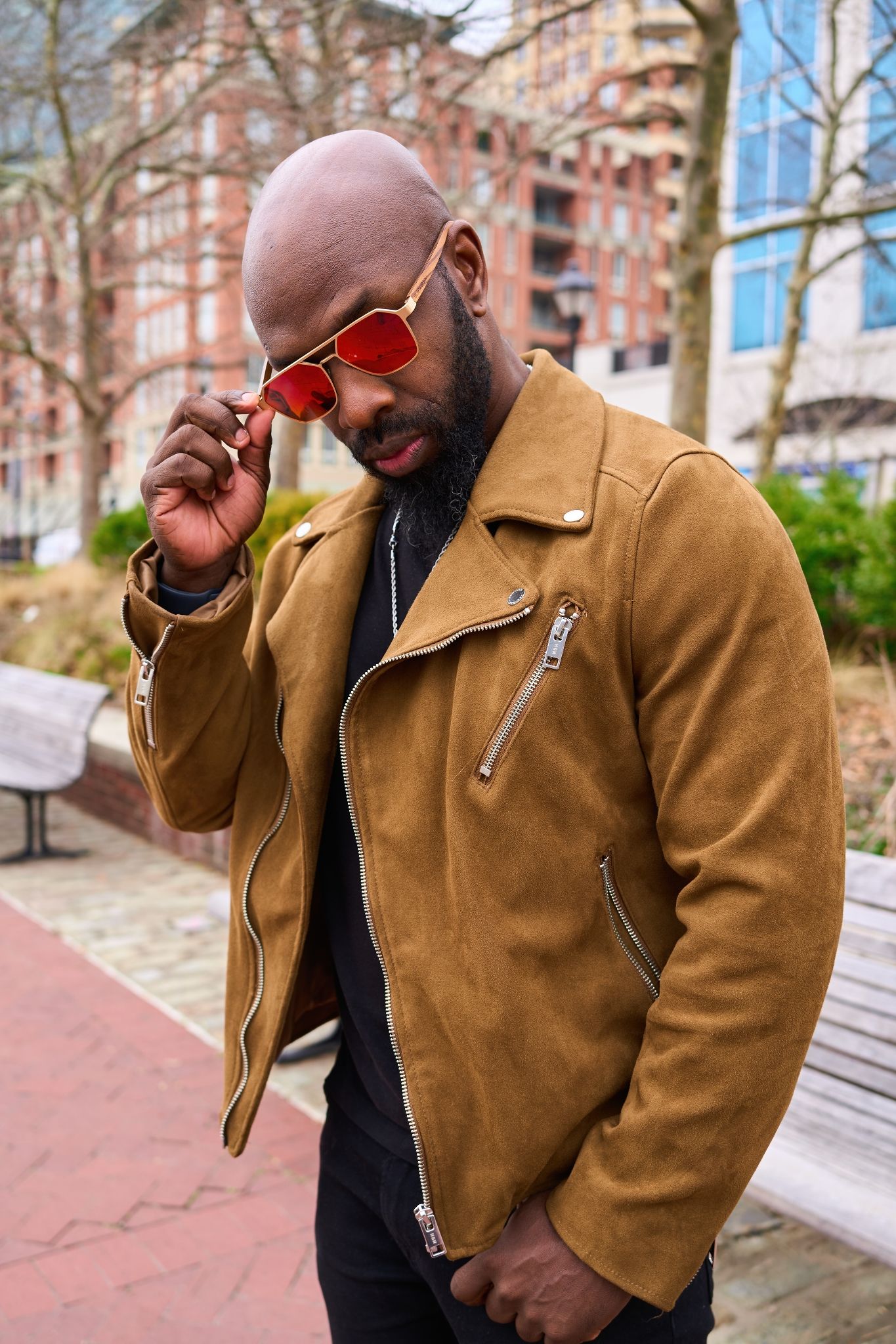 Stylish person in tan suede jacket and red sunglasses poses confidently on urban walkway.