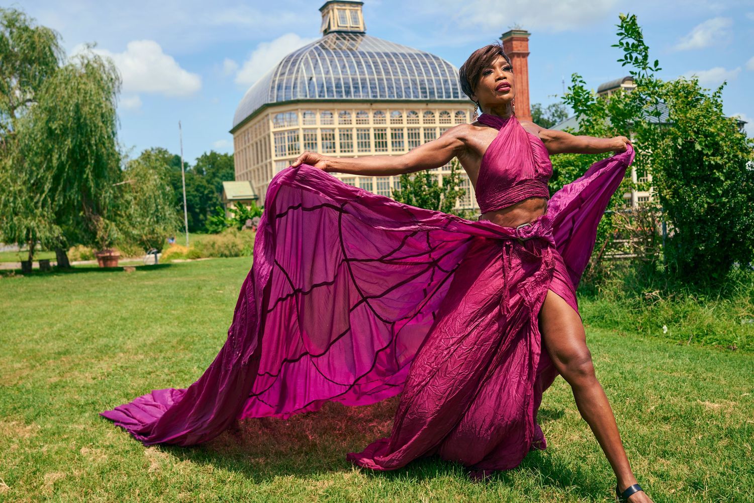 Woman twirling in vibrant purple skirt set in lush garden with greenhouse in background.
