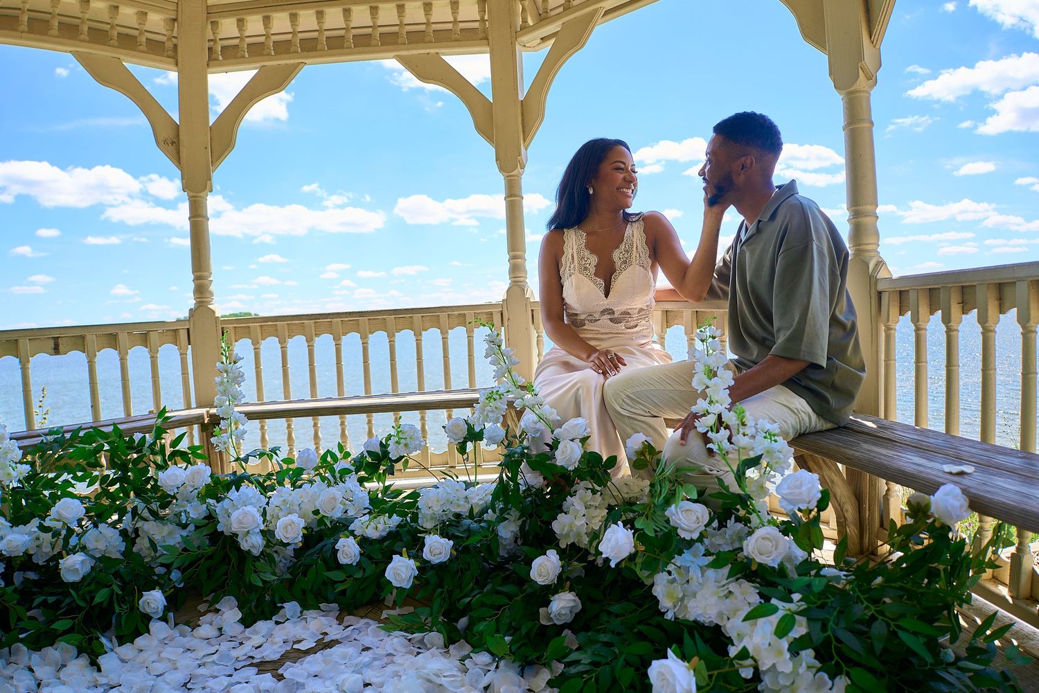 Happy couple sharing an intimate moment seated in a white gazebo adorned with lush white flowers under a bright blue sky.