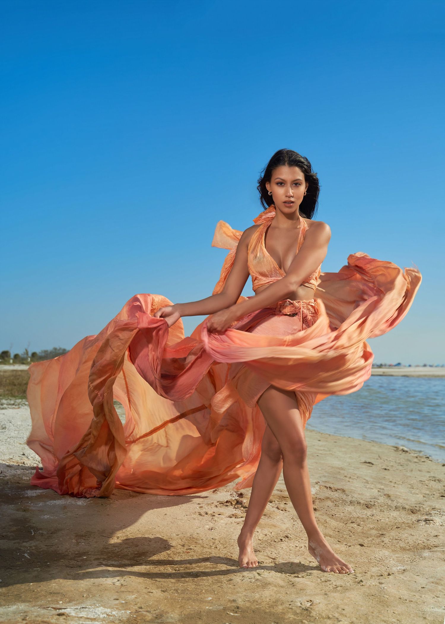Flowing peach dress catches wind dramatically on sunny beach shoreline.